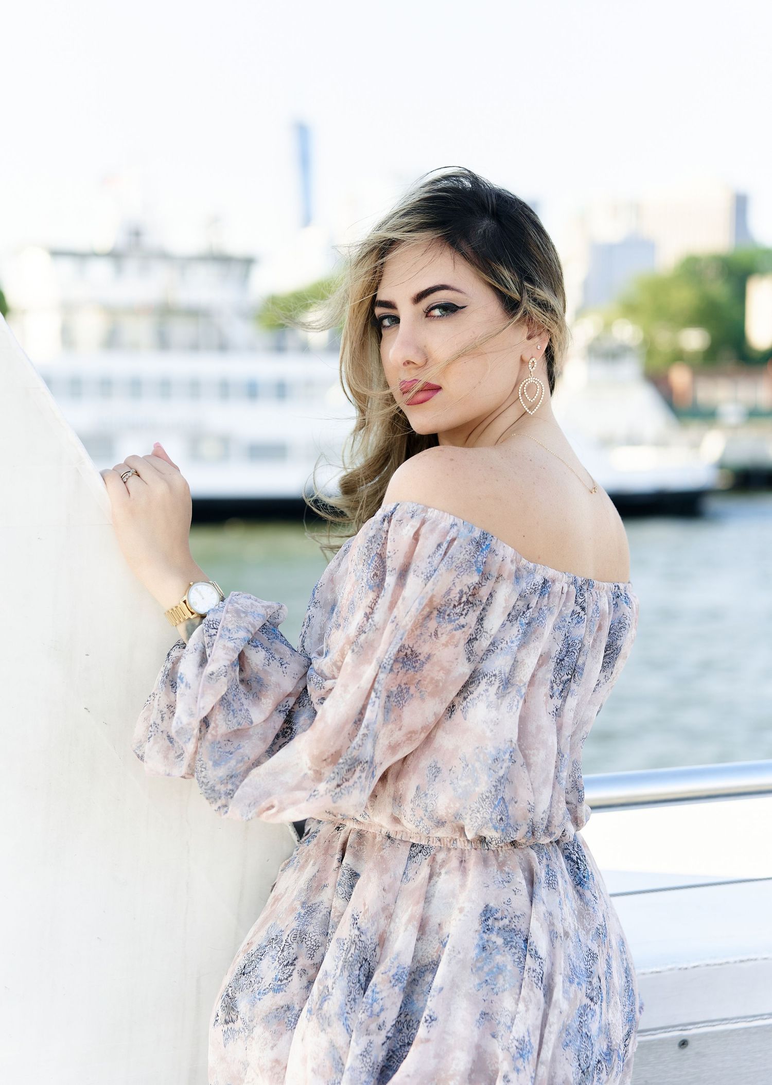 Woman in floral off-shoulder dress posing near waterfront railing with city skyline in background.