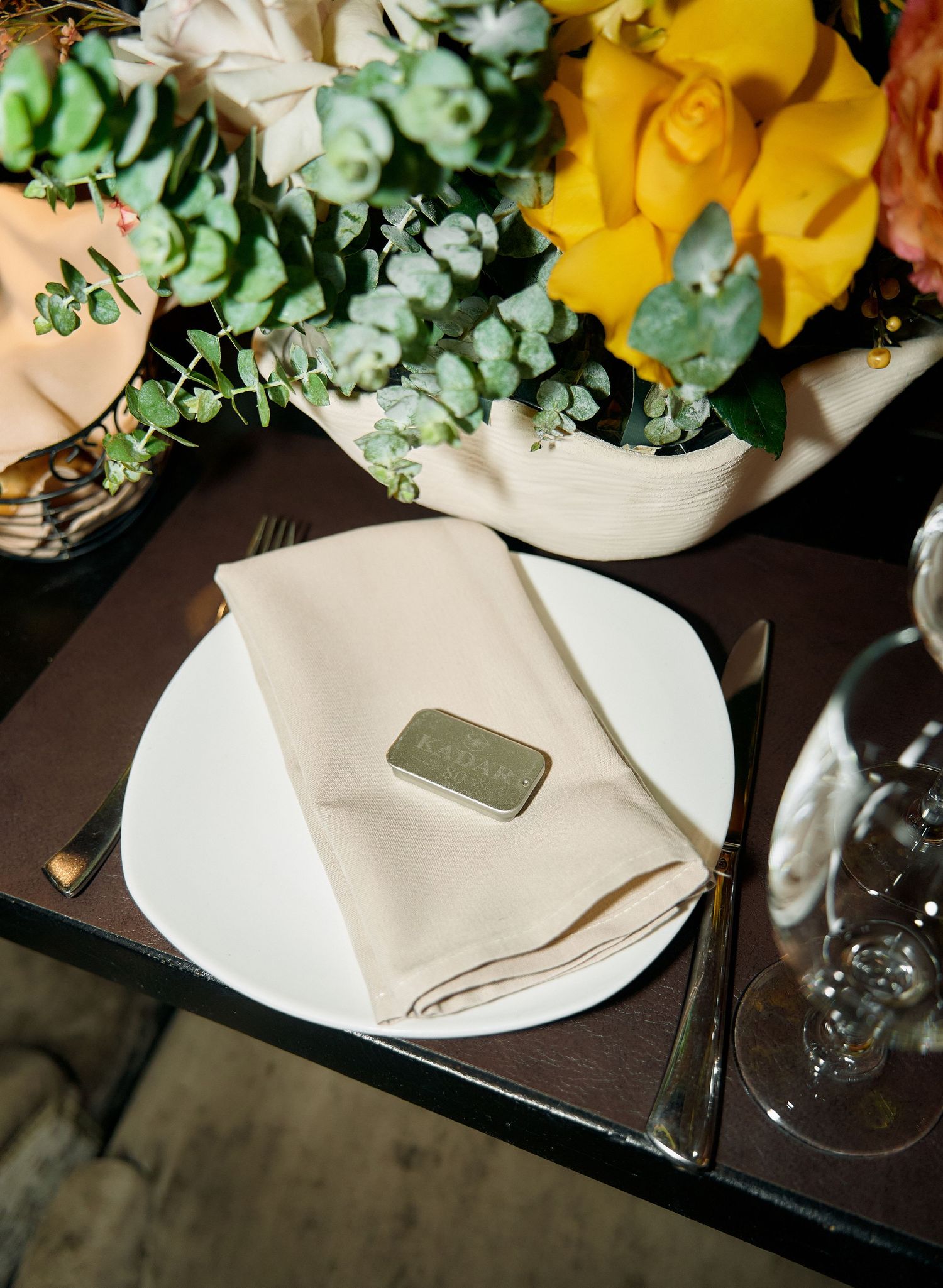 Elegant table setting with beige linen napkin, place card, gold cutlery, and yellow floral centerpiece.