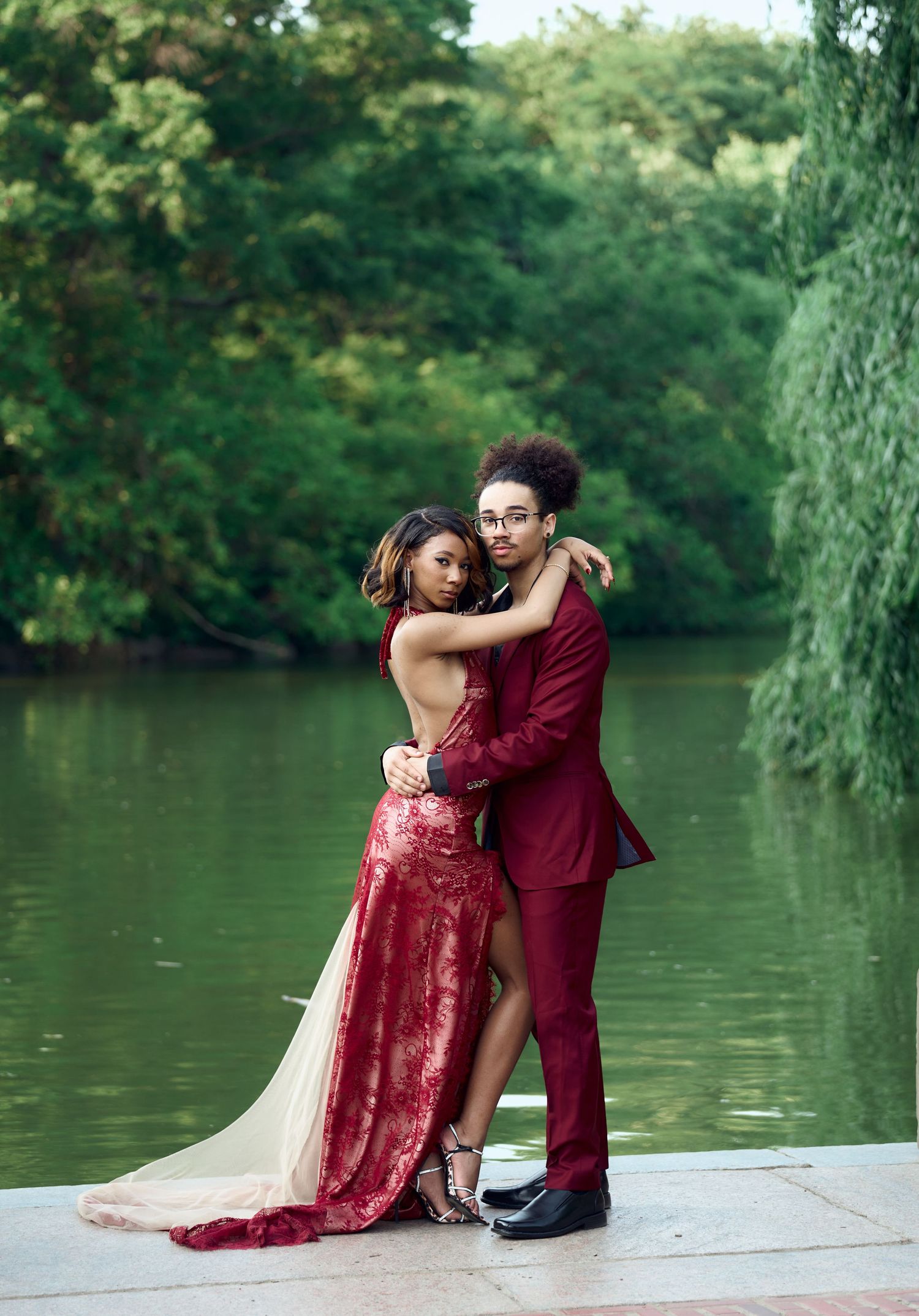 Couple in coordinating red lace dress and burgundy suit pose embracing by a lakeside.