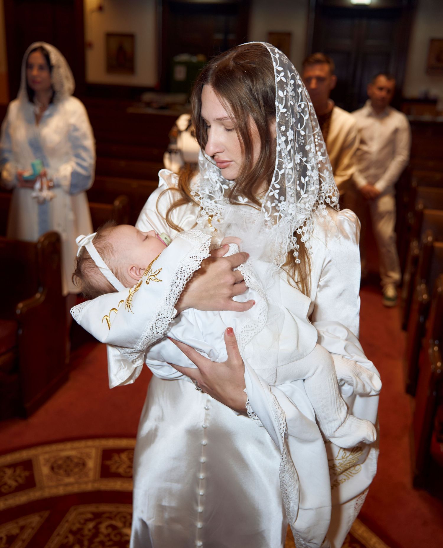 Woman in white lace veil holding a sleeping baby dressed in white during a christening ceremony.