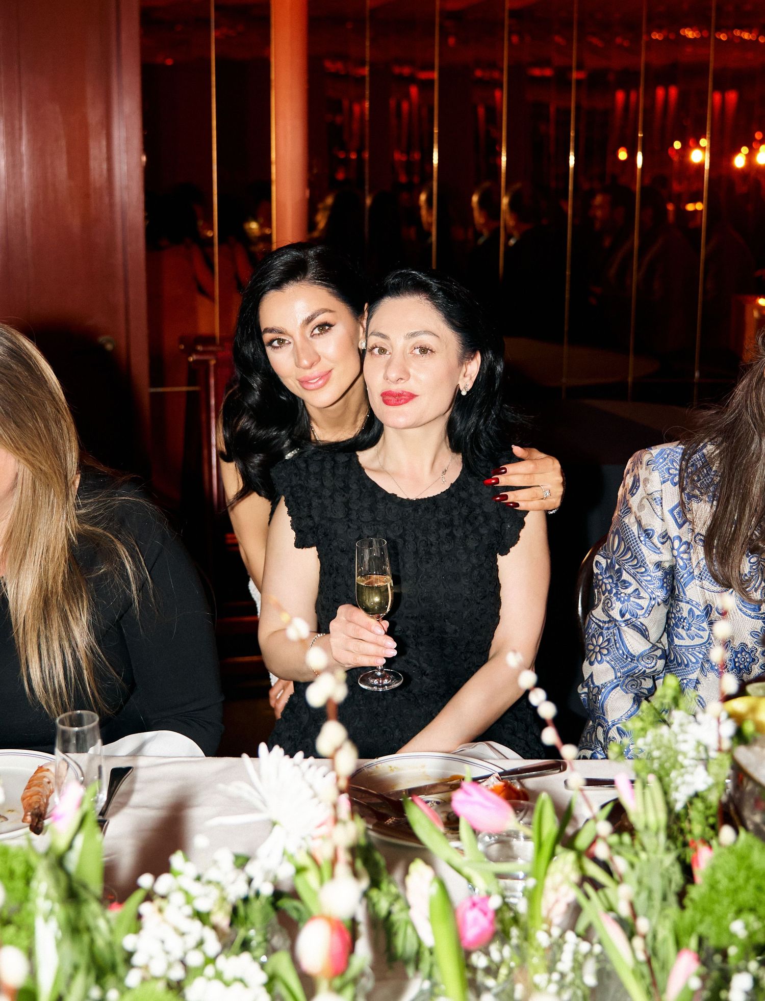 Two women in black dresses posing together at a restaurant table, one holding a champagne glass.
