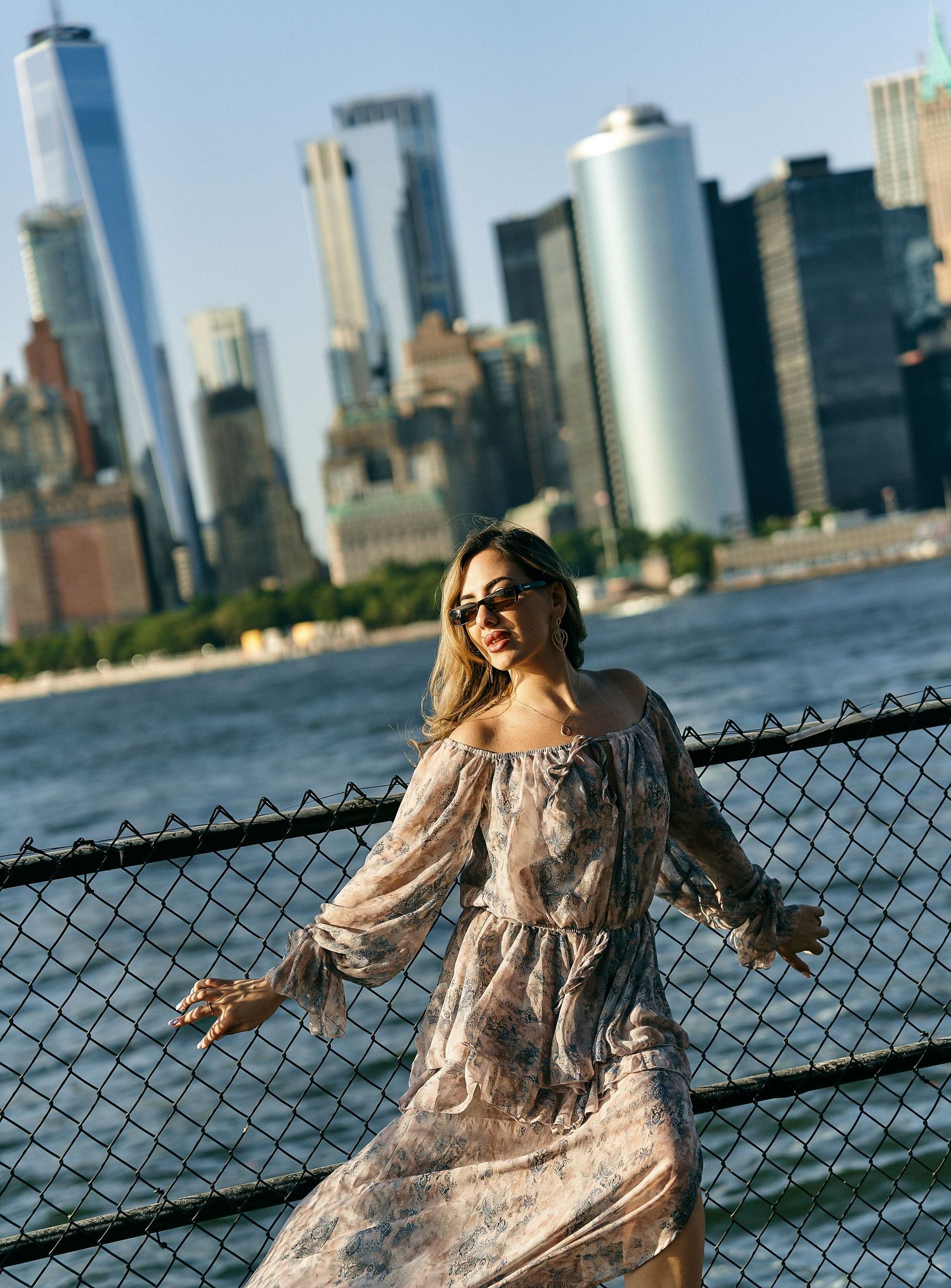 Woman in floral off-shoulder dress wearing sunglasses near NYC skyline.