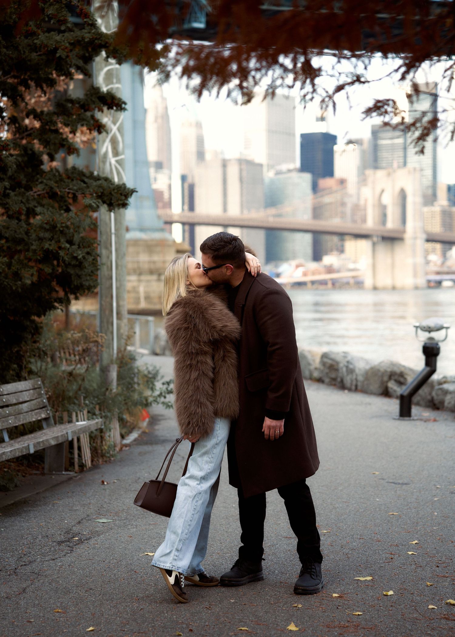 Couple sharing a kiss on a waterfront path with the Brooklyn Bridge and NYC skyline behind them.