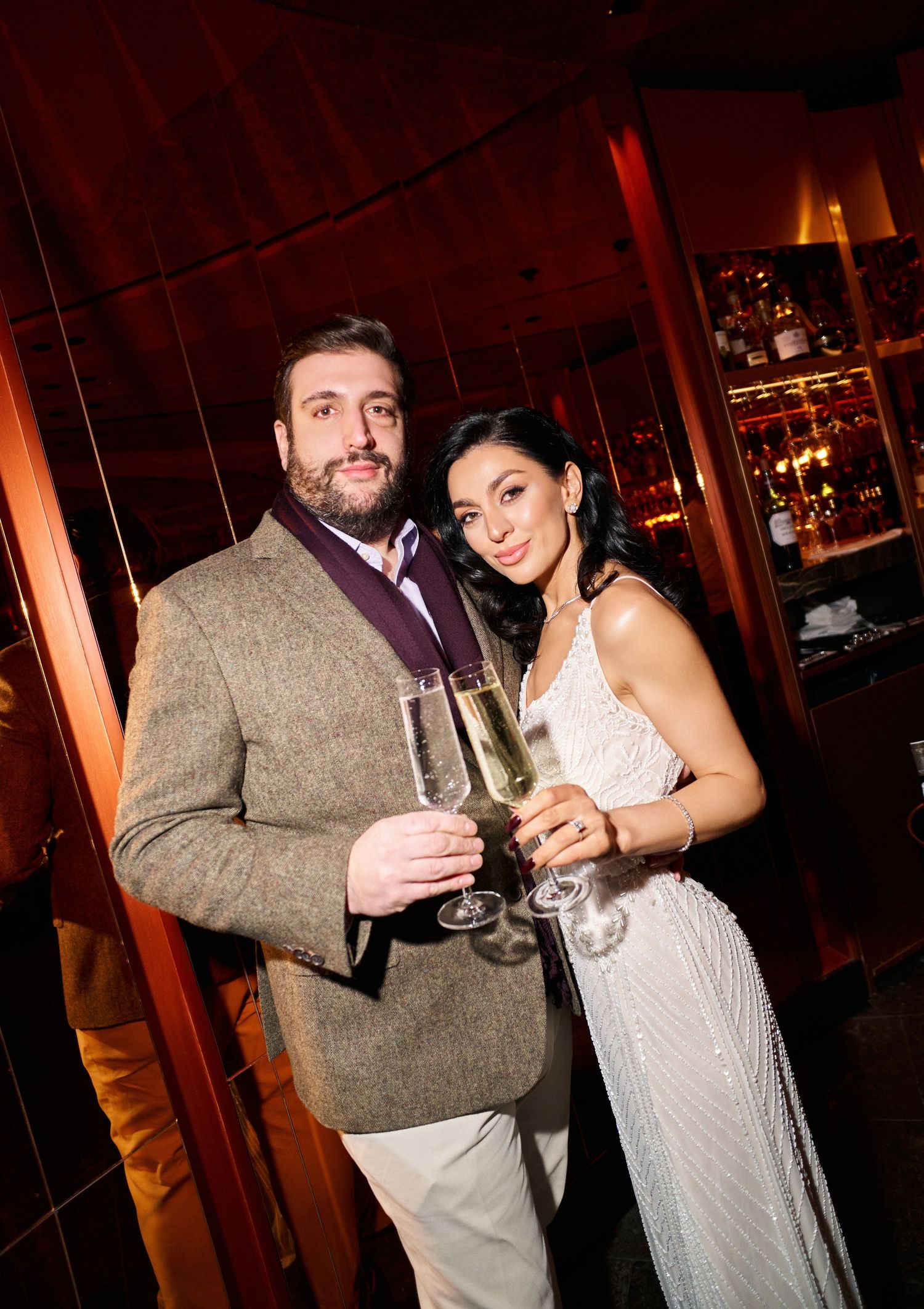 Smiling couple toasting with champagne flutes at an upscale bar, woman in beaded gown, man in blazer.
