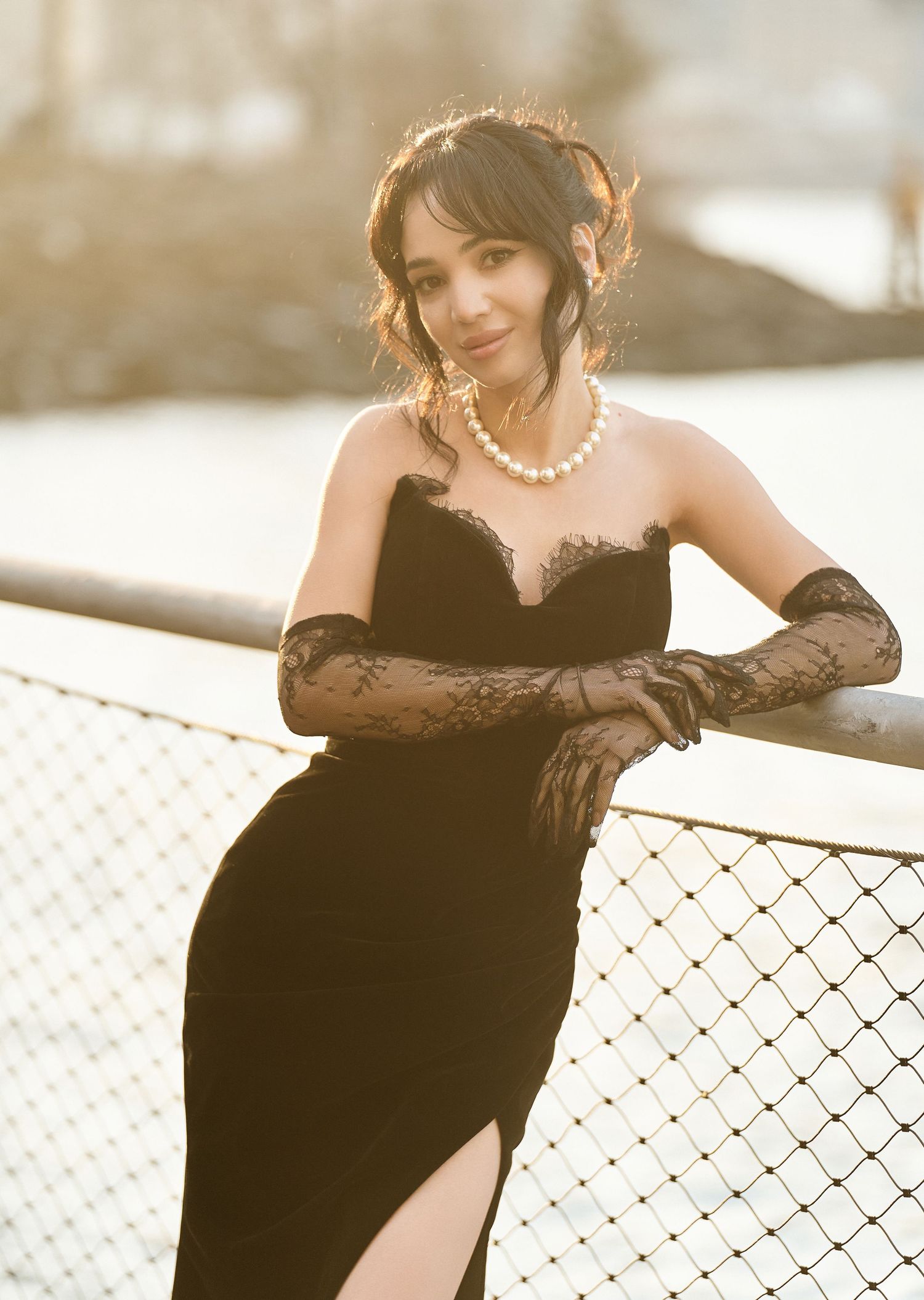 Elegant woman in black velvet gown with lace gloves and pearl necklace leans on railing outdoors.