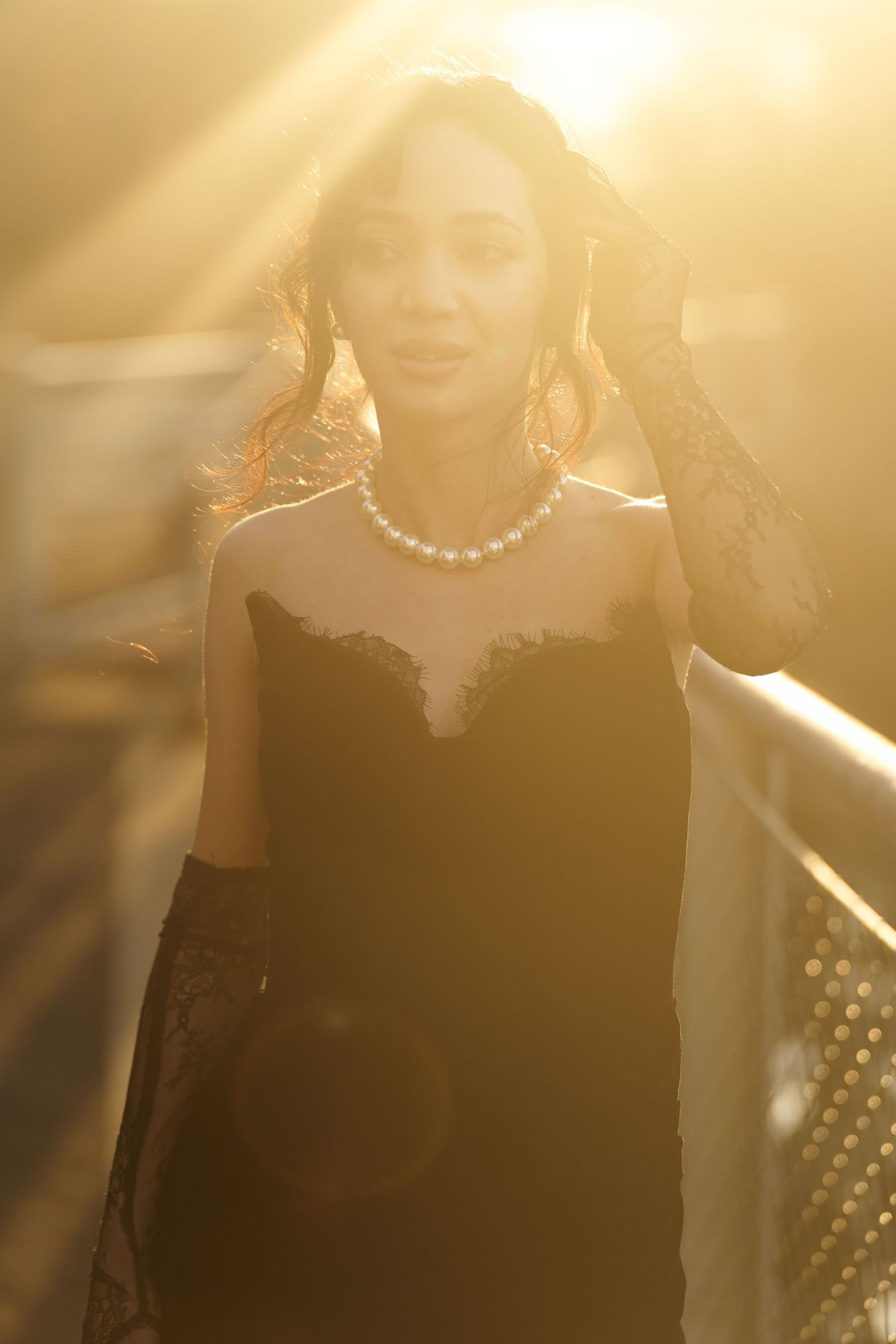 Elegant woman in black lace gown with pearl necklace stands in golden backlight.