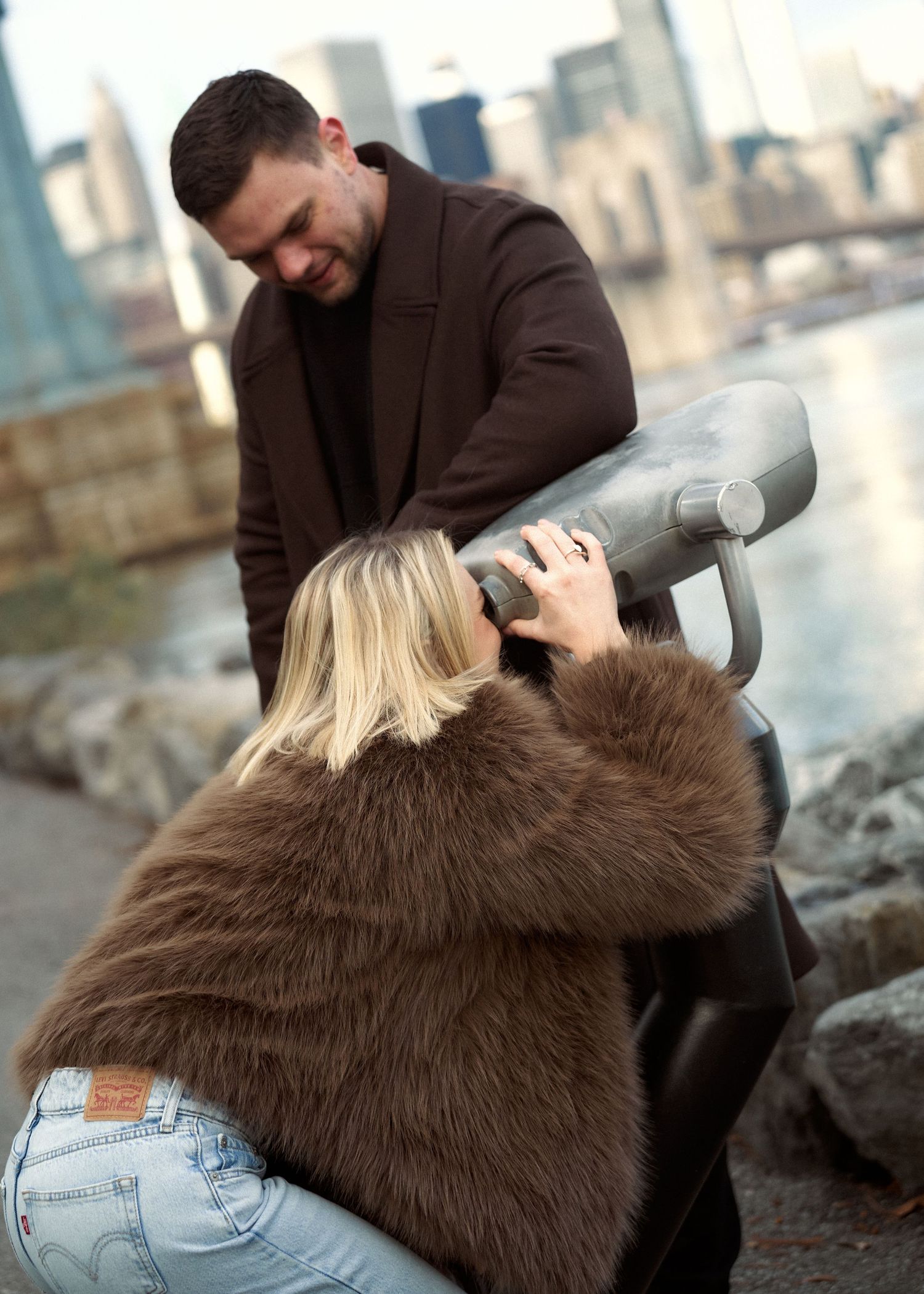 Blonde woman in fur coat using a coin-operated viewfinder while man in brown coat watches nearby.