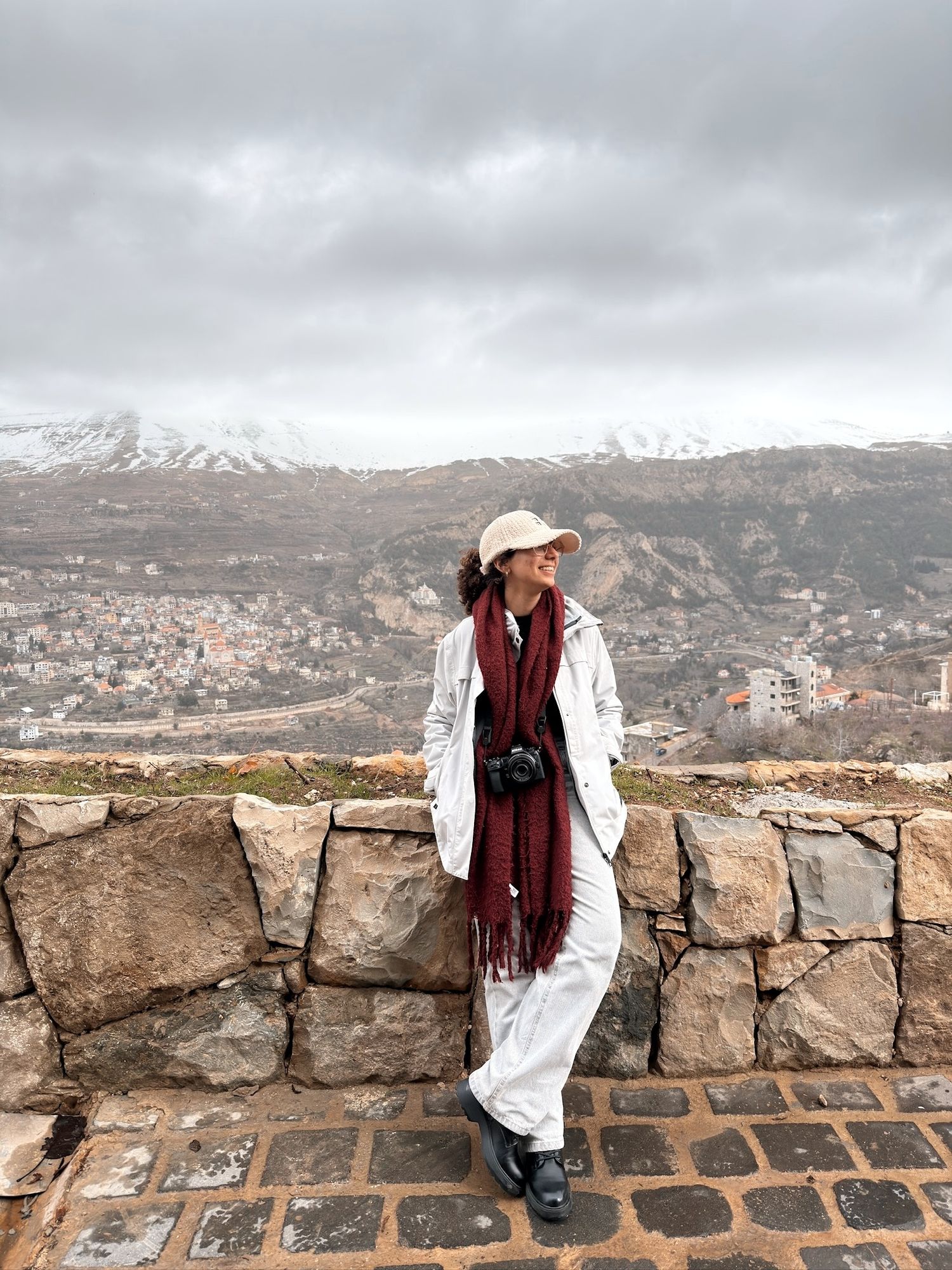 Person in white outfit and red scarf leaning on stone wall with snowy mountain town backdrop on cloudy day.