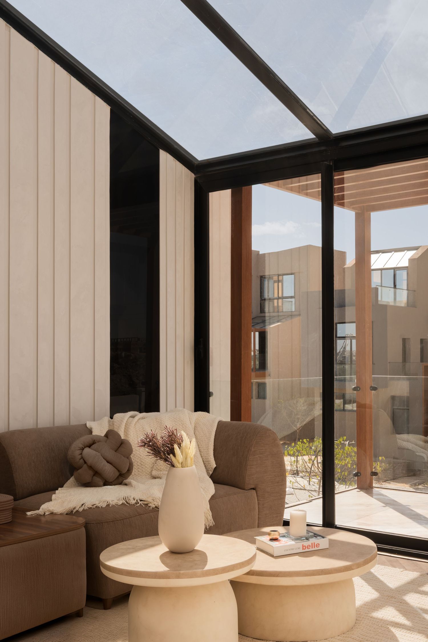 Modern sunroom with glass roof, beige sofa, round coffee tables, and vase of dried flowers overlooking a courtyard.
