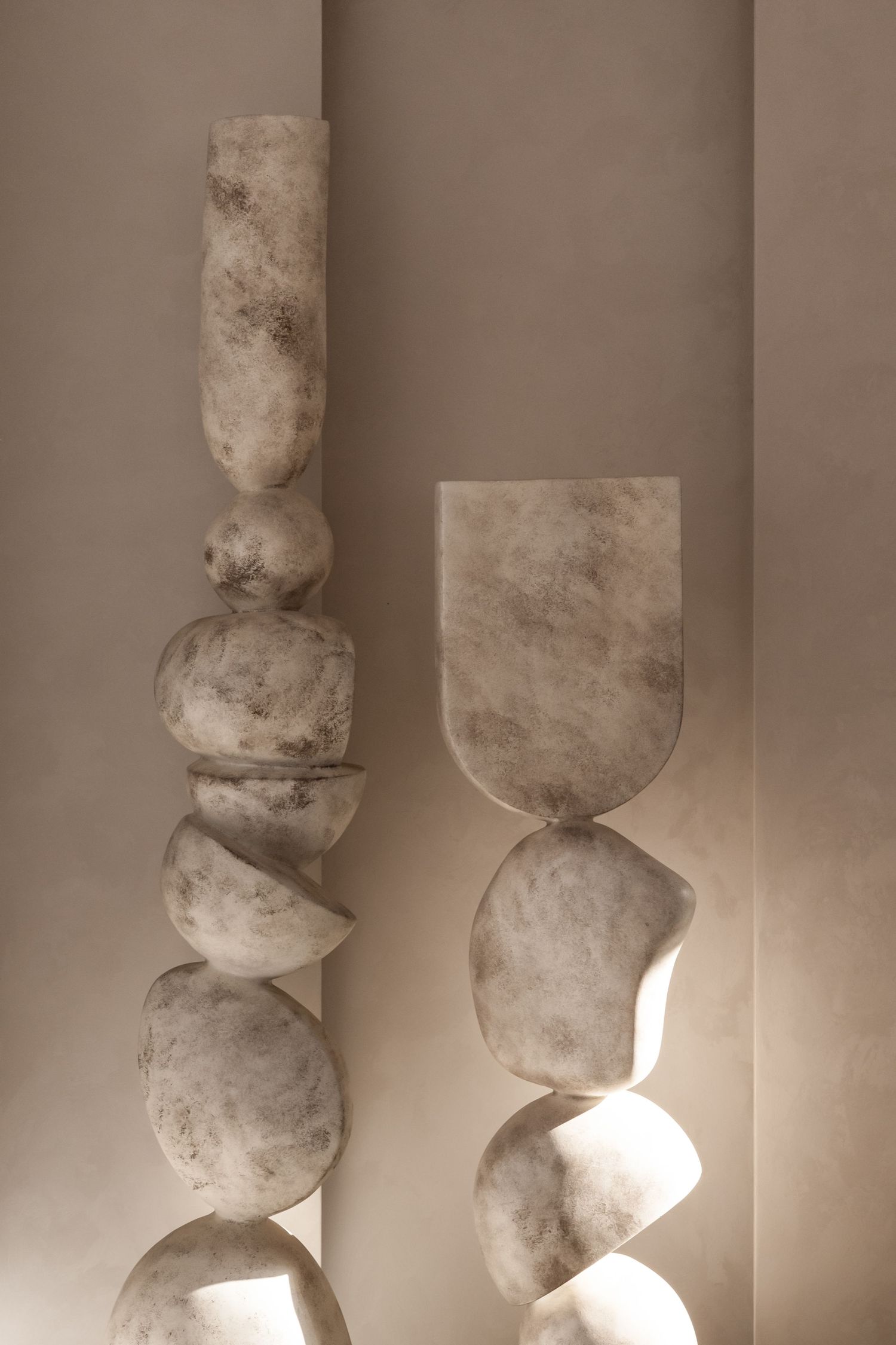 Two abstract marble sculptures with stacked organic stone forms stand against a warm beige wall in soft natural light.