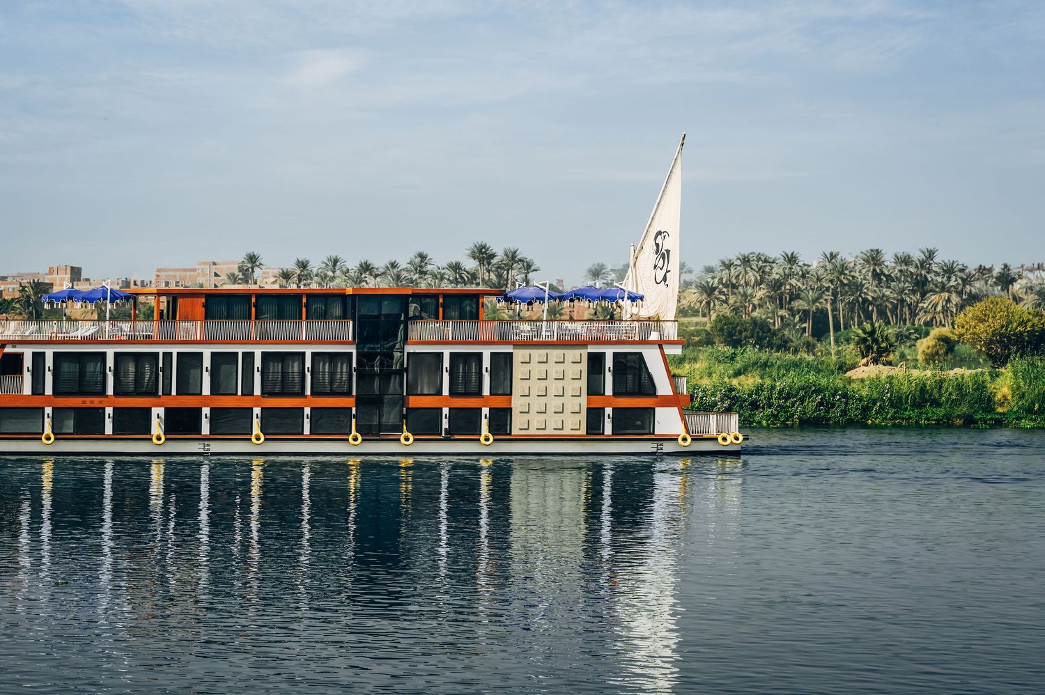 A panoramic view of a waterfront building with palm trees and boats reflected in calm waters on a sunny day.