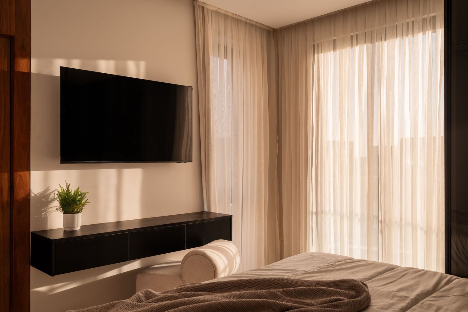 Modern bedroom with wall-mounted TV, floating shelf, potted plant, and sheer curtains by a sunlit window.