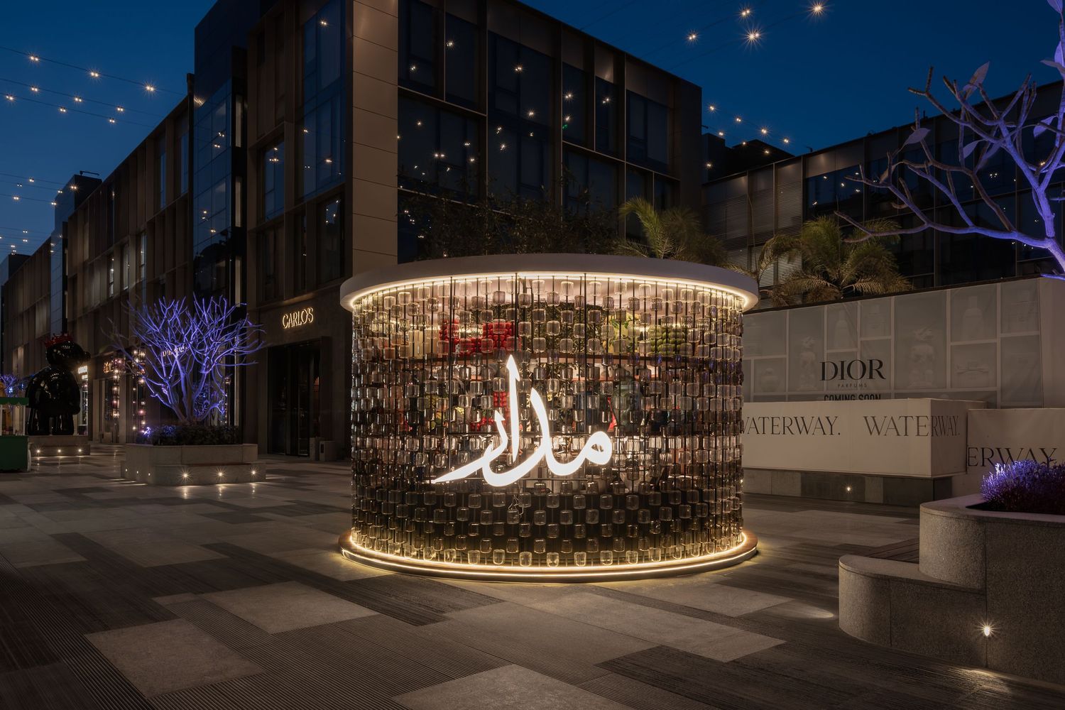 Illuminated Arabic calligraphy kiosk glows at night near Dior store at Waterway retail center.
