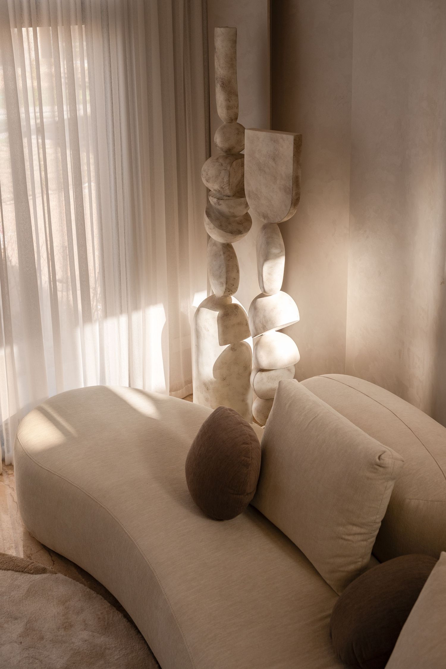 Minimalist living room with cream curved sofa, brown pillow, and abstract stone sculpture near sheer curtains.