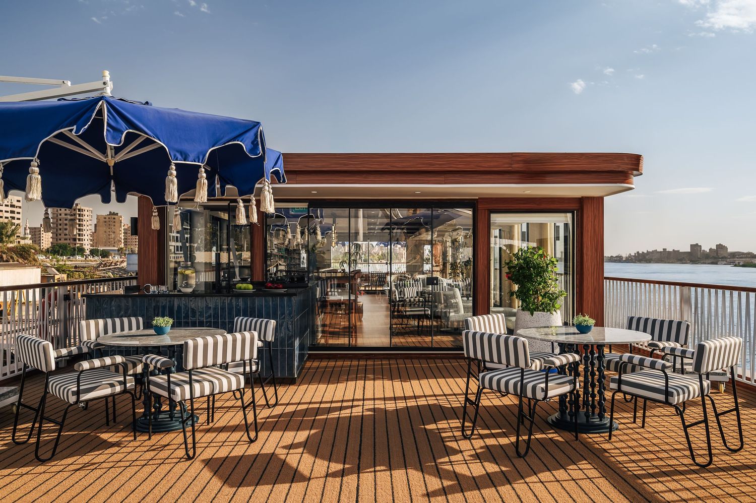 Outdoor deck with dining tables and blue umbrellas overlooking waterfront. Interior Photography