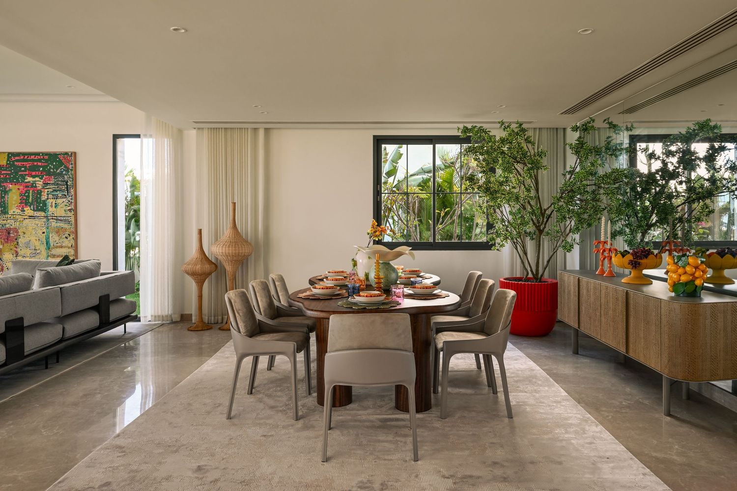 Modern dining room with round wooden table, gray chairs, lush plants, and natural light from large windows.