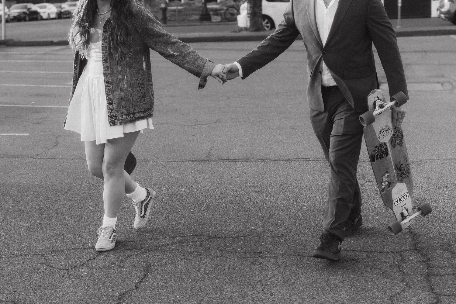 A couple holds hands while carrying skateboards in a parking lot, captured in black and white.