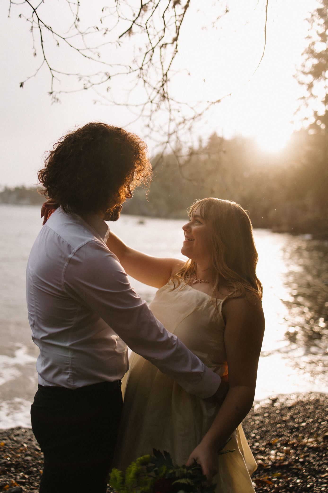A romantic couple embraces by a lake at sunset, with soft golden light filtering through trees in the background.