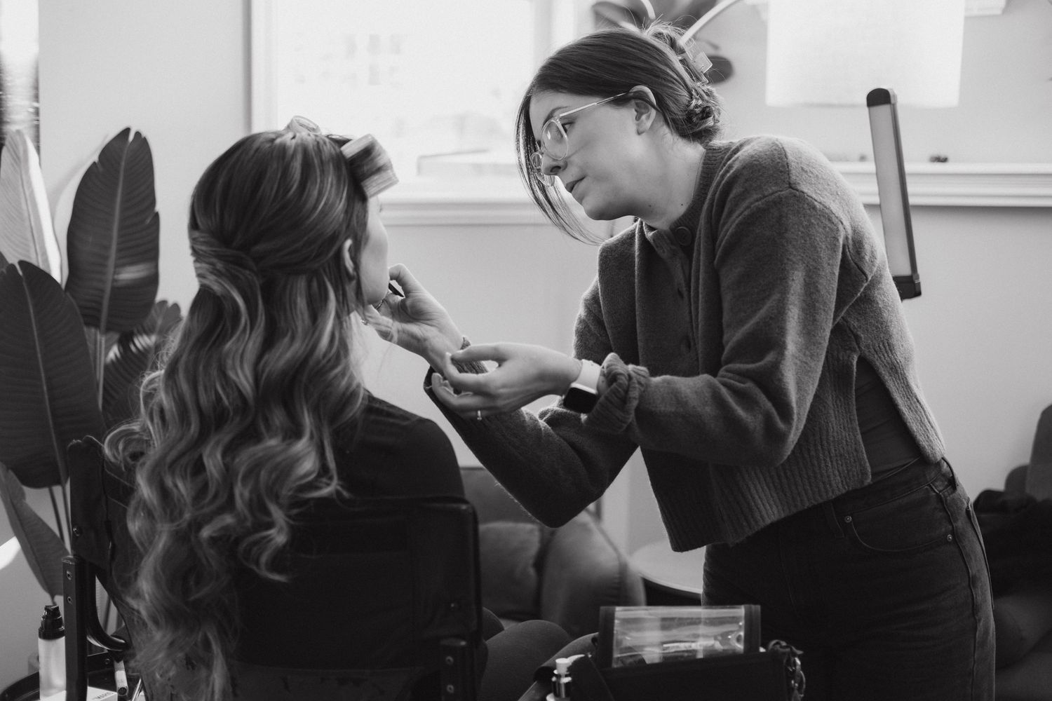Professional makeup artist applies cosmetics to a client with long wavy hair in a black and white photograph.