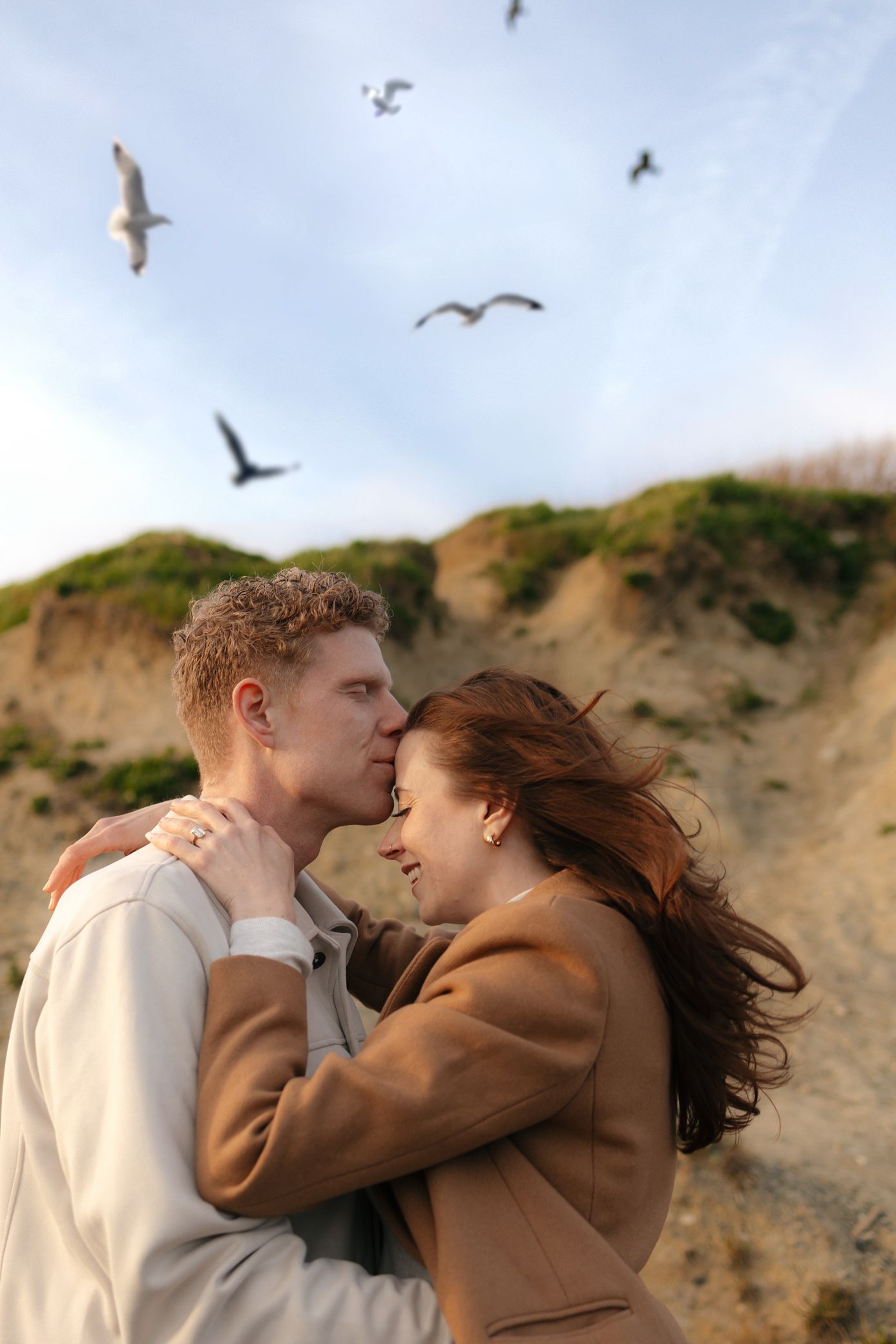 A couple embracing on a cliff with seagulls flying overhead.