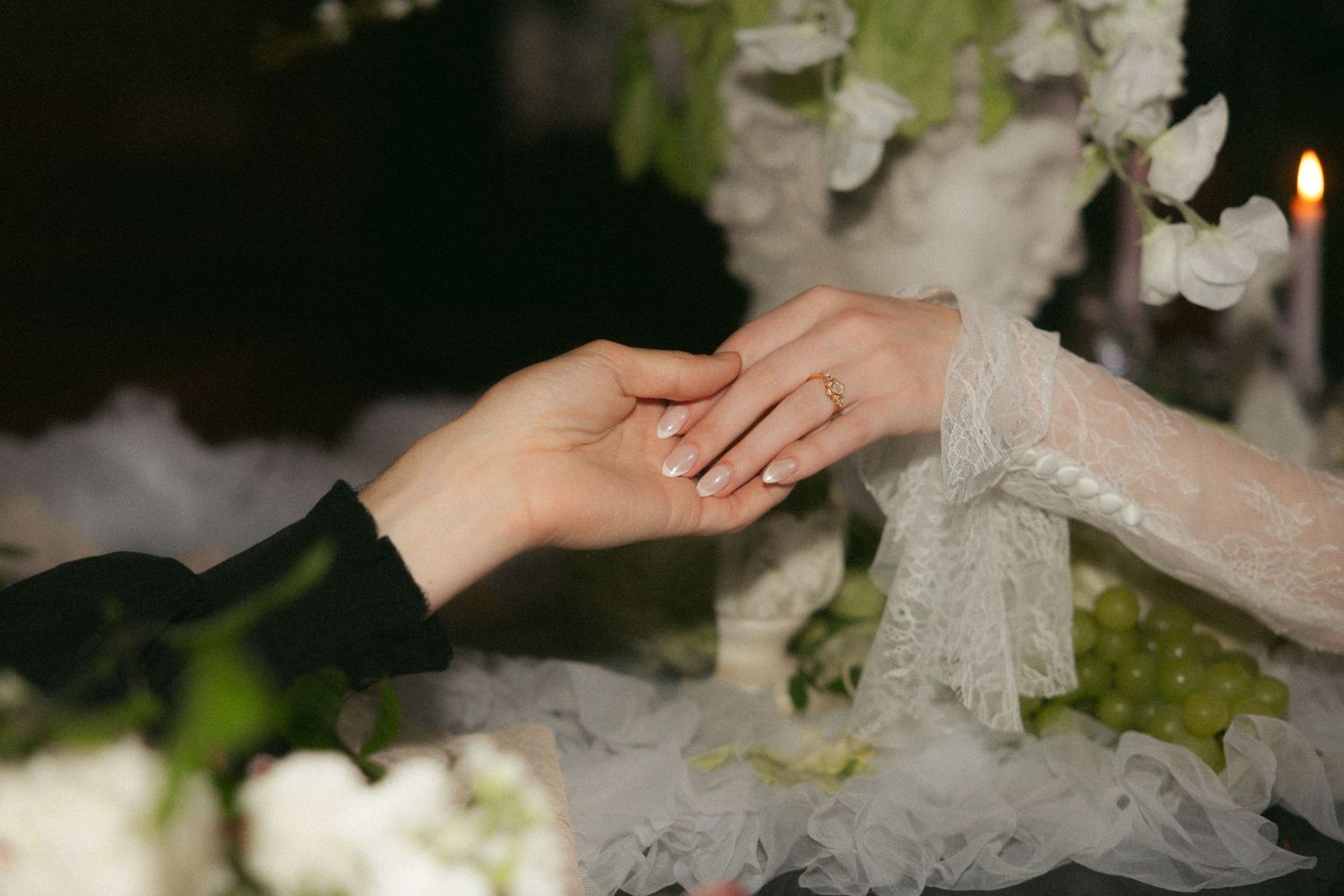 Close-up of couple exchanging rings during wedding ceremony with white floral decor and candlelight.