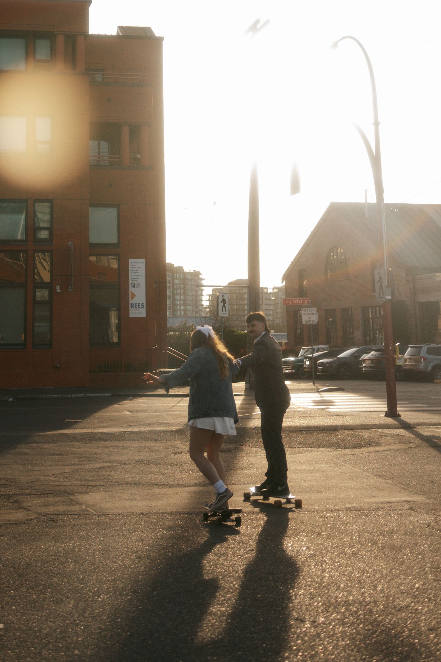 Two people skateboarding together on an empty street at sunset with long shadows cast on the pavement.
