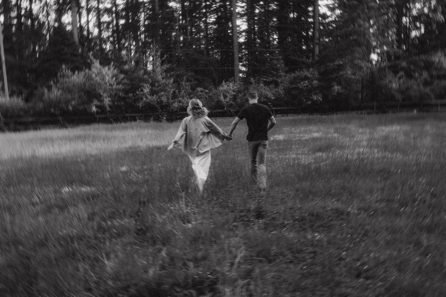 A couple runs through a misty meadow surrounded by tall evergreen trees at sunset, captured in black and white.
