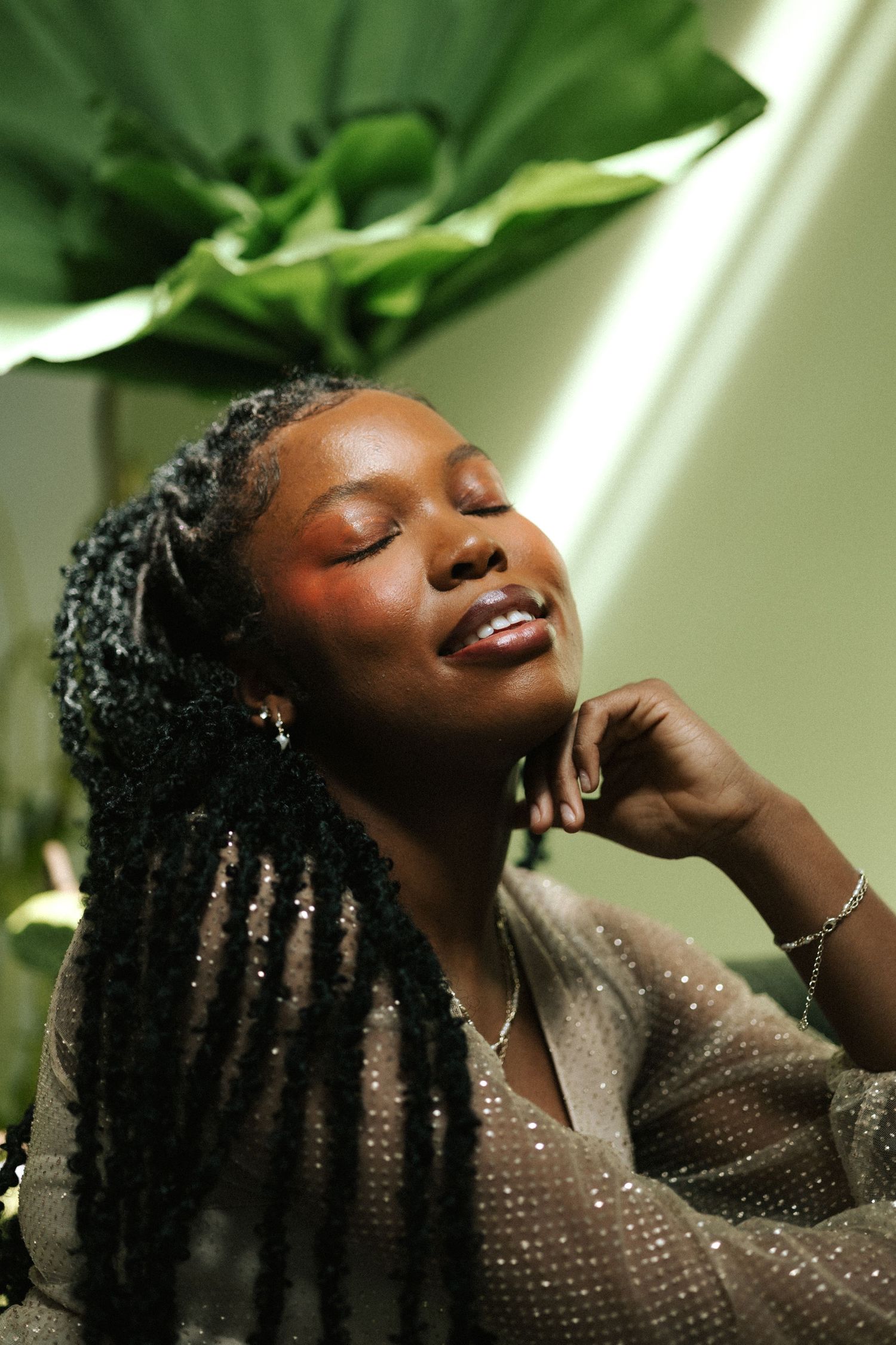 A young Black woman with braids poses eyes closed, wearing a shimmery top with rosy blush makeup near tropical plants.