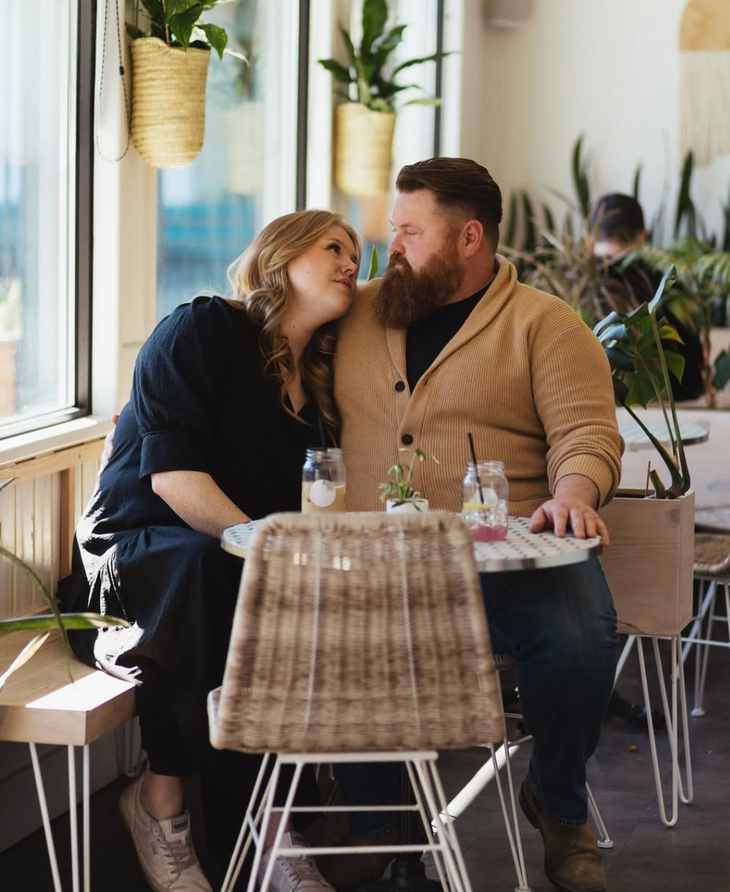 Candid cafe scene of couple sharing tender moment by window with hanging plants.