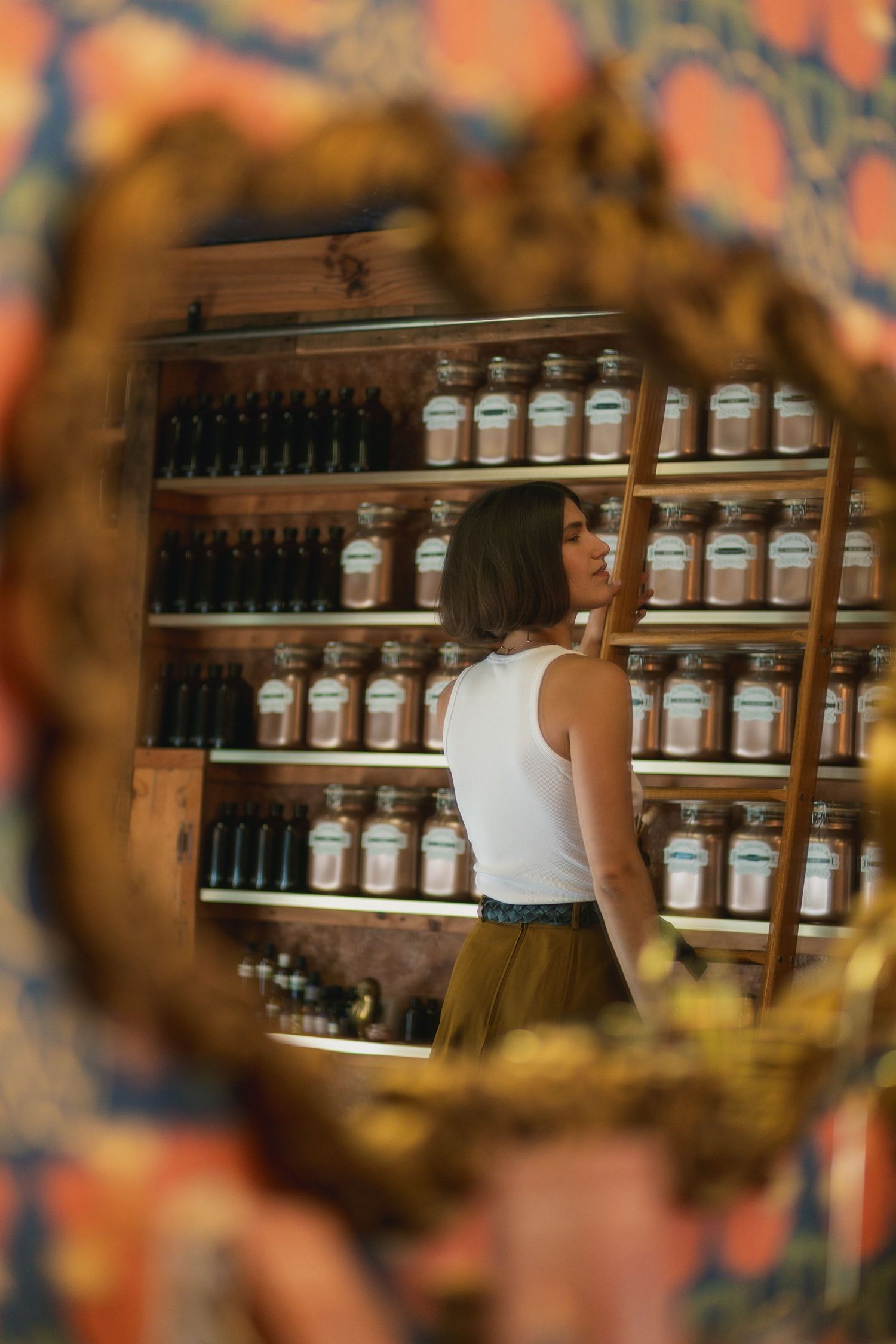 Woman in white tank top browsing shelves of apothecary jars in a boutique shop, reflected in ornate gold mirror.
