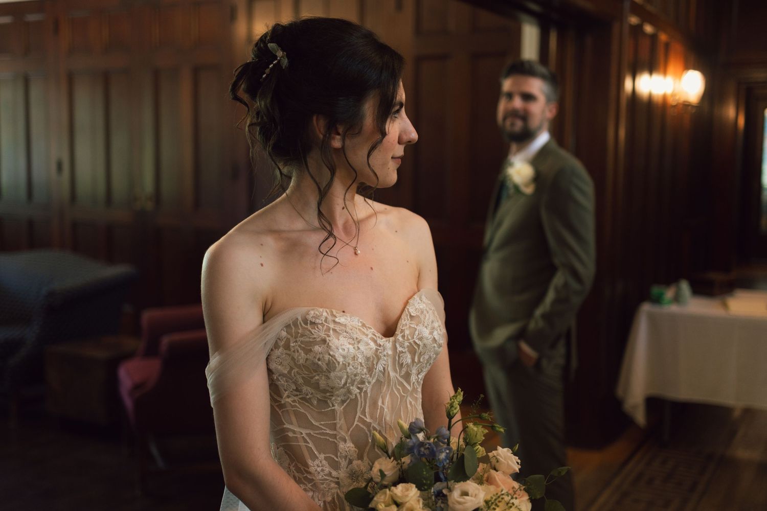 Bride in strapless lace gown holds yellow bouquet while groom stands in background at dimly lit venue.