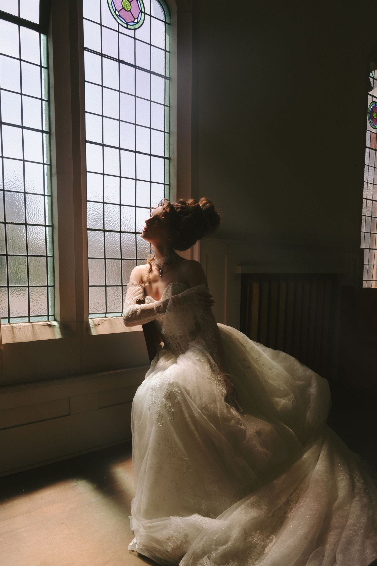 Bride in a flowing white tulle wedding gown standing by leaded glass windows in soft natural light.