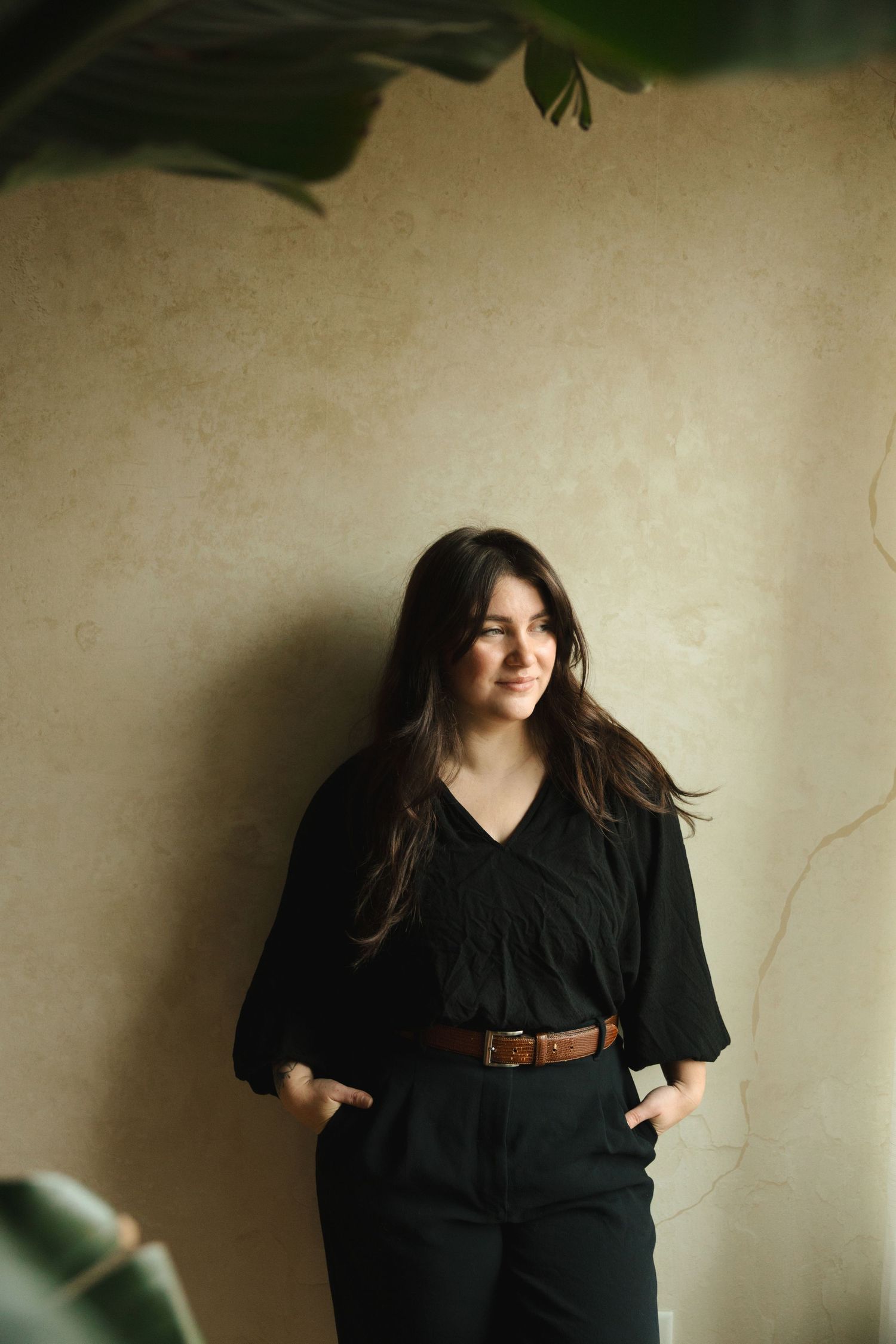 A person in a black outfit with brown belt stands against a beige wall with soft lighting and plant shadows.