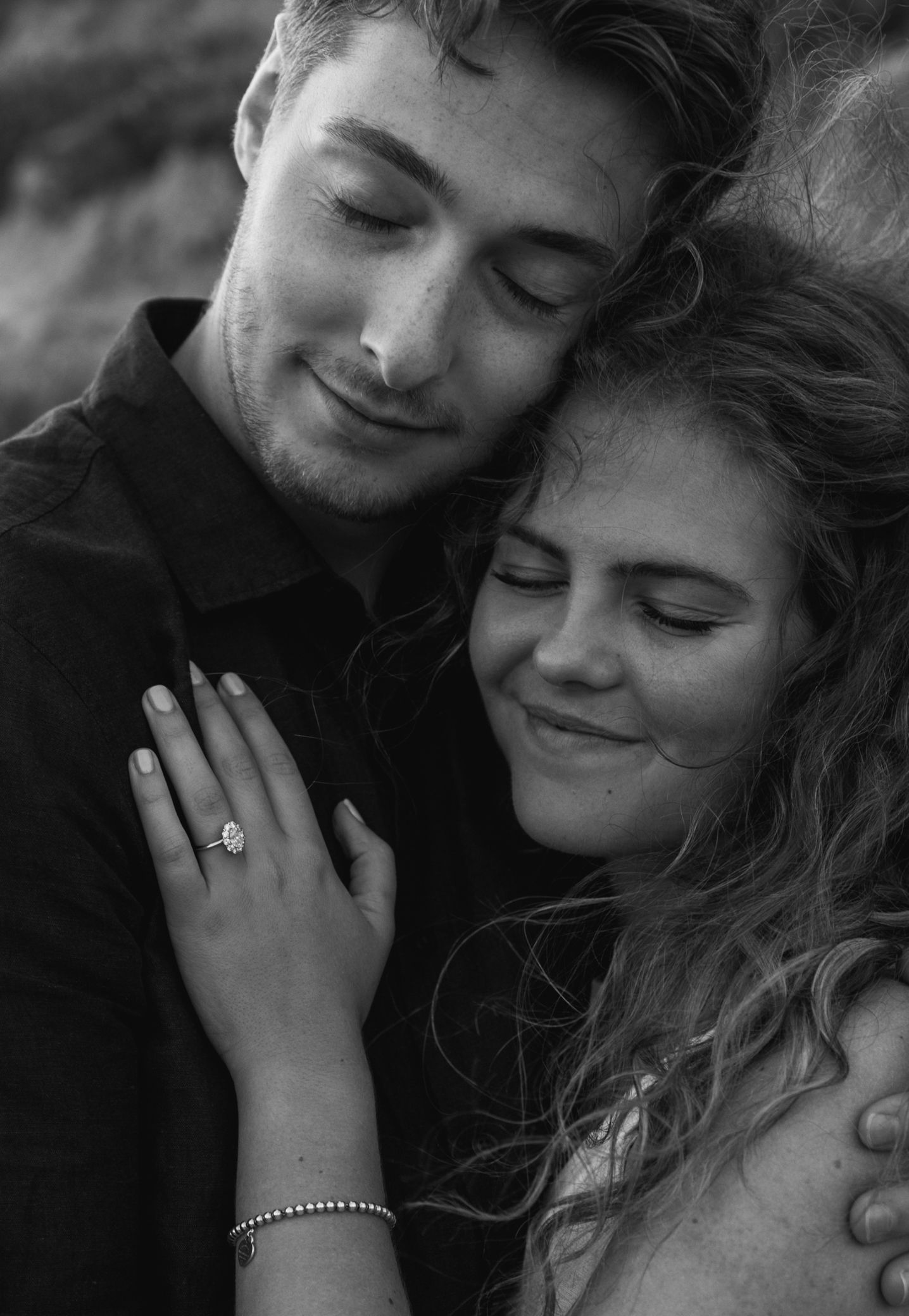Black and white photo of a romantic couple embracing, woman wearing an engagement ring, eyes closed, sharing a tender moment.