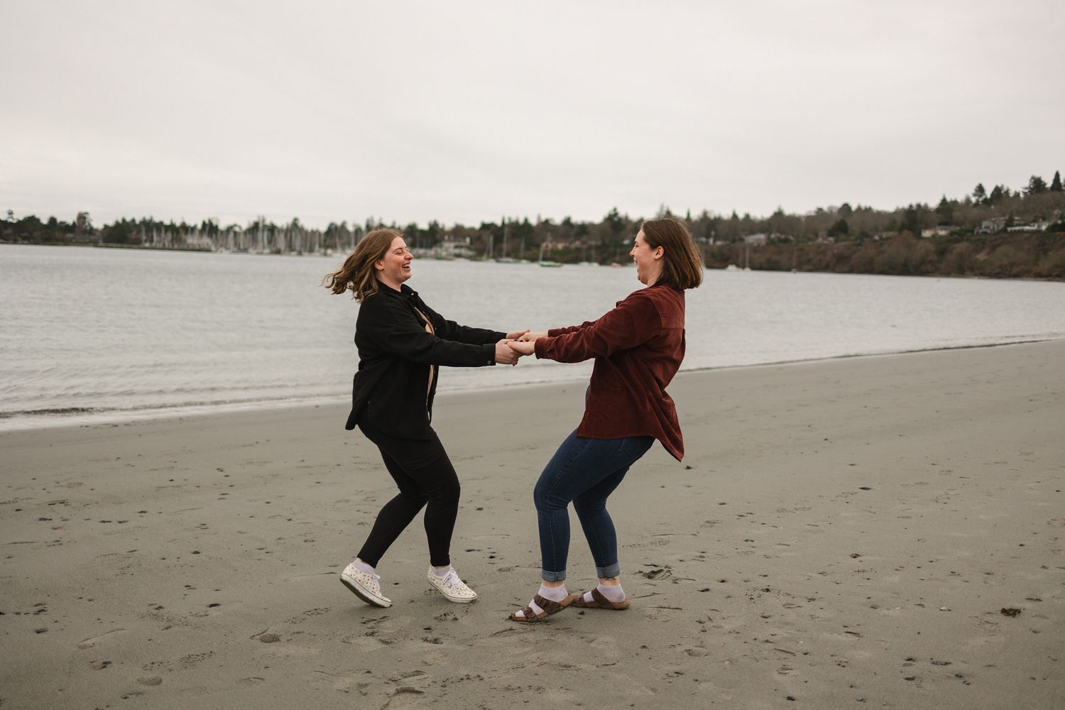 Two people hold hands while dancing and spinning together on a cloudy beach with water and trees in the background.