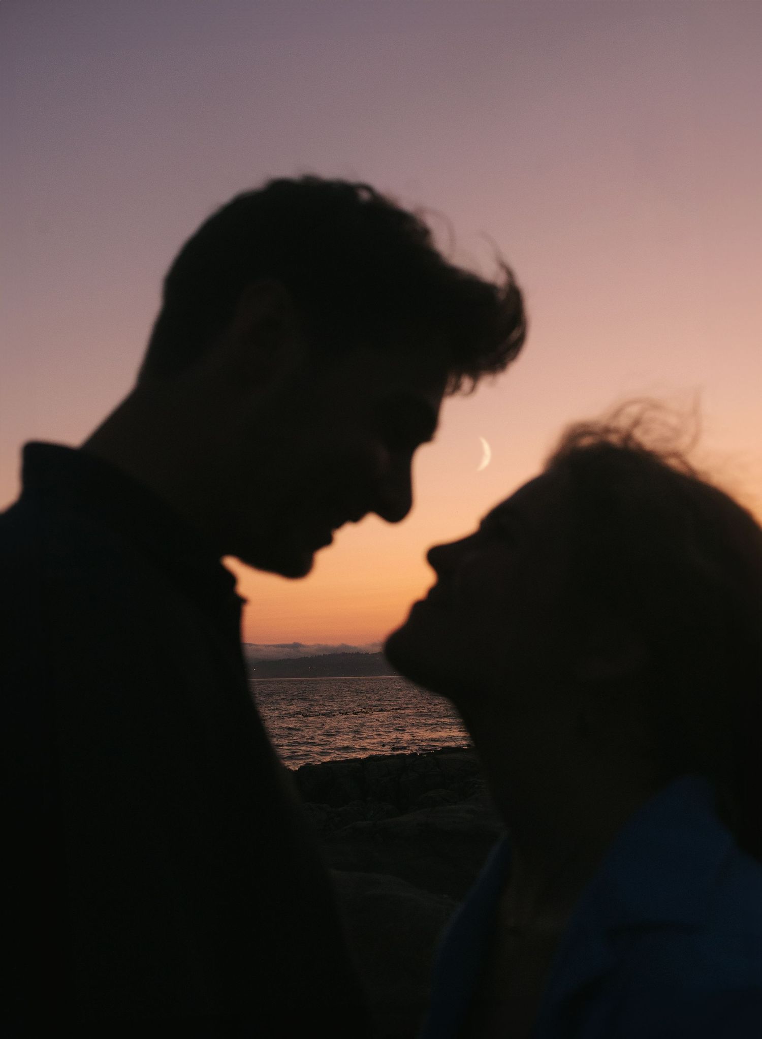 Romantic silhouette of a couple about to kiss at sunset by the ocean with a crescent moon visible.