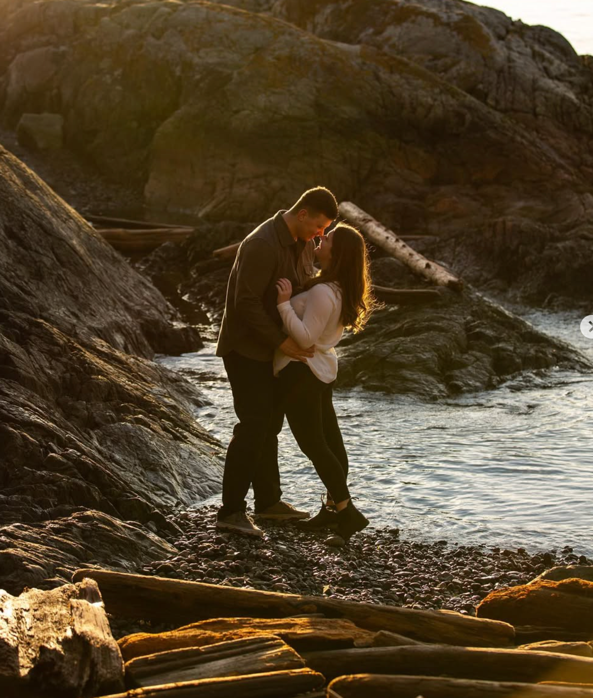Couple embracing on rocky coastline during golden hour with ocean waves in background.