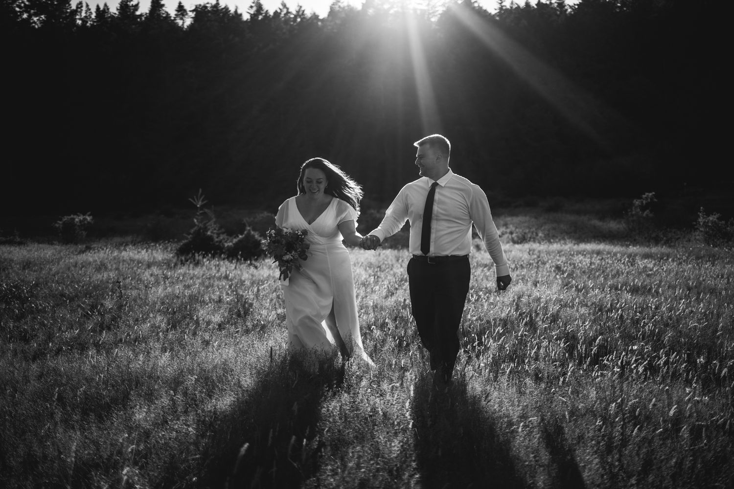 A couple walks through a sunlit meadow with dramatic sun flare behind trees.