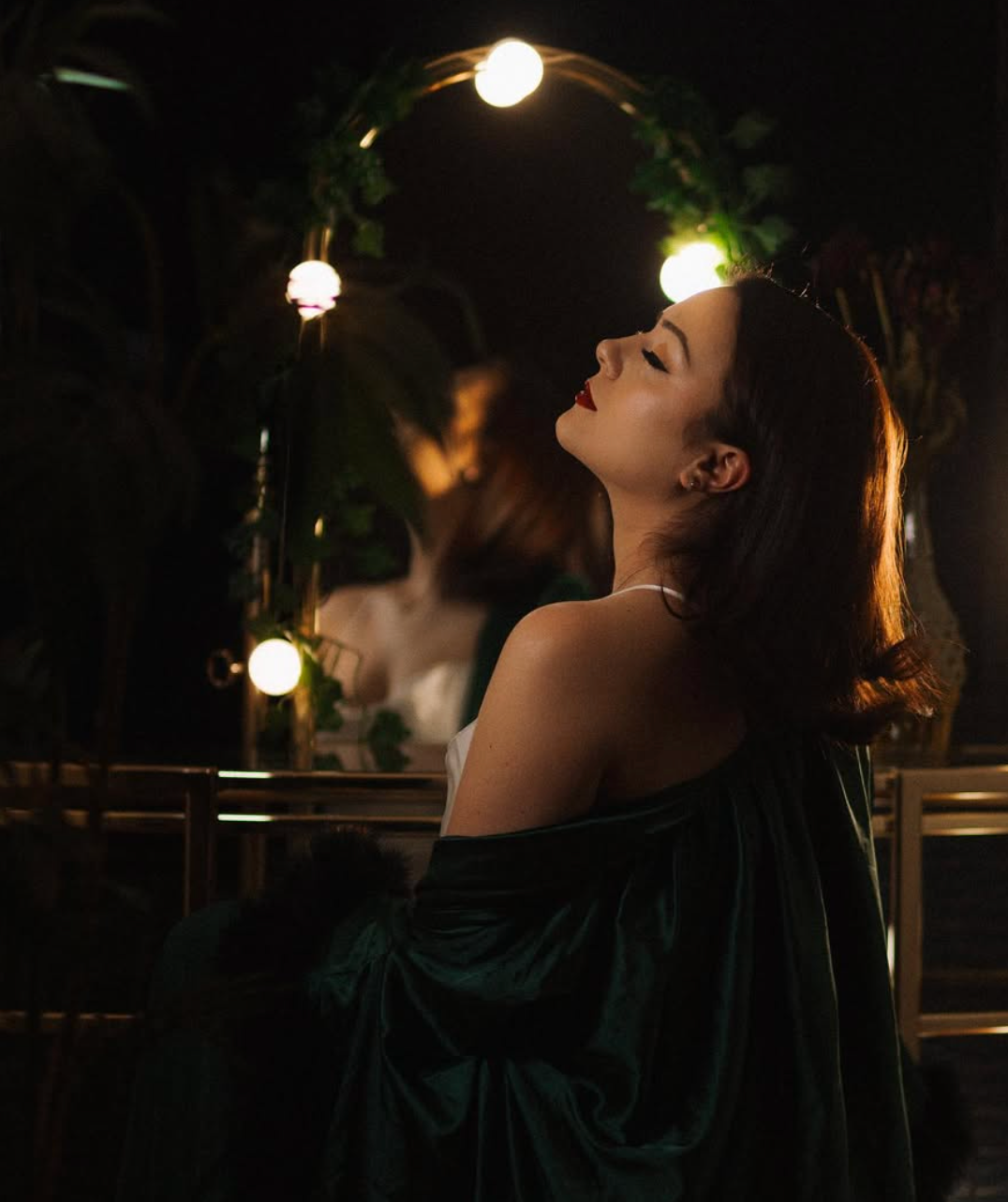 Artistic nighttime portrait with glowing string lights illuminating a draped emerald green dress in a garden setting.