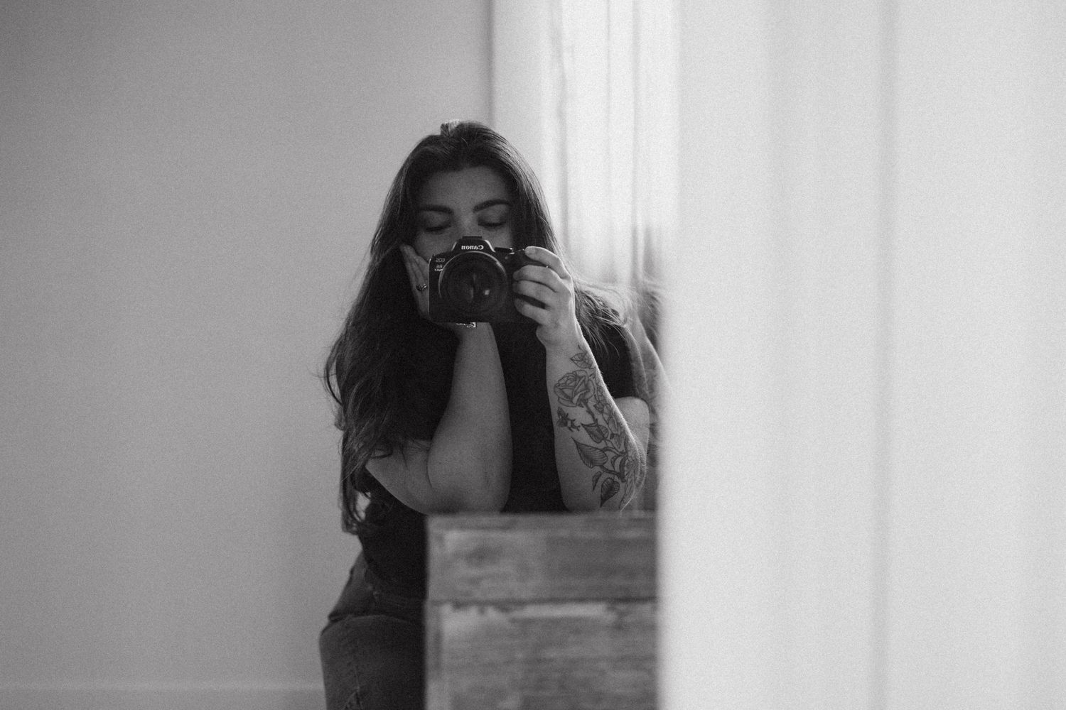 Black and white photo of a tattooed woman taking a mirror selfie with a DSLR camera indoors.