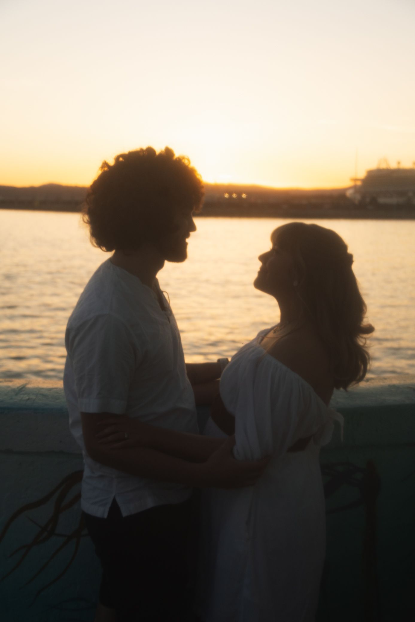 Silhouette of a couple embracing by the waterfront during sunset with soft golden light reflecting on the water.