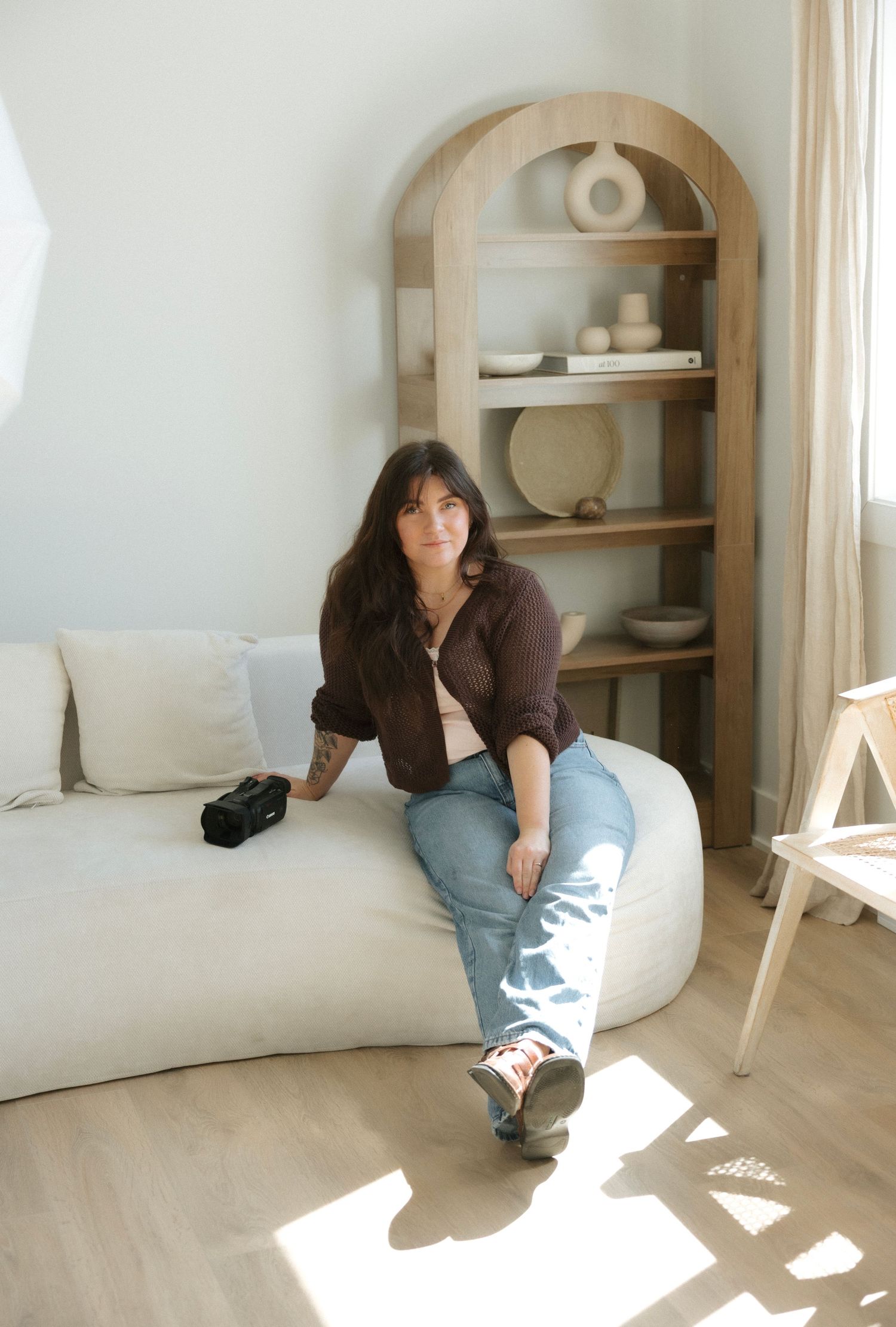 Woman in brown shirt and jeans sits on white sofa near wooden arched shelf in bright minimalist room.