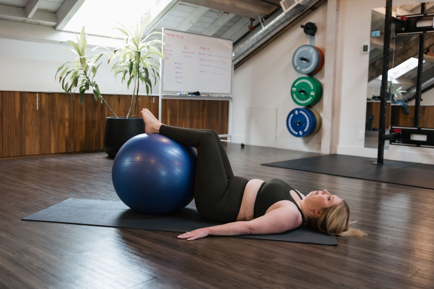 Person demonstrating proper plank form on a yoga mat in a fitness studio with exercise balls and wooden floors.