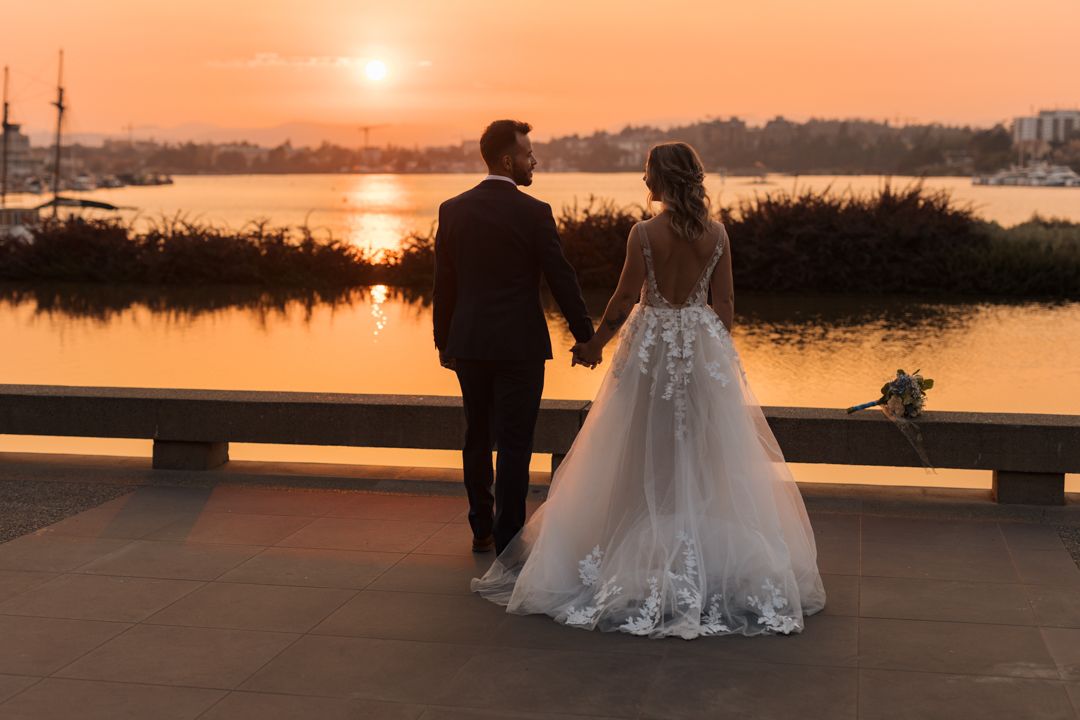 A newlywed couple admiring a sunset over water while wearing elegant wedding attire.