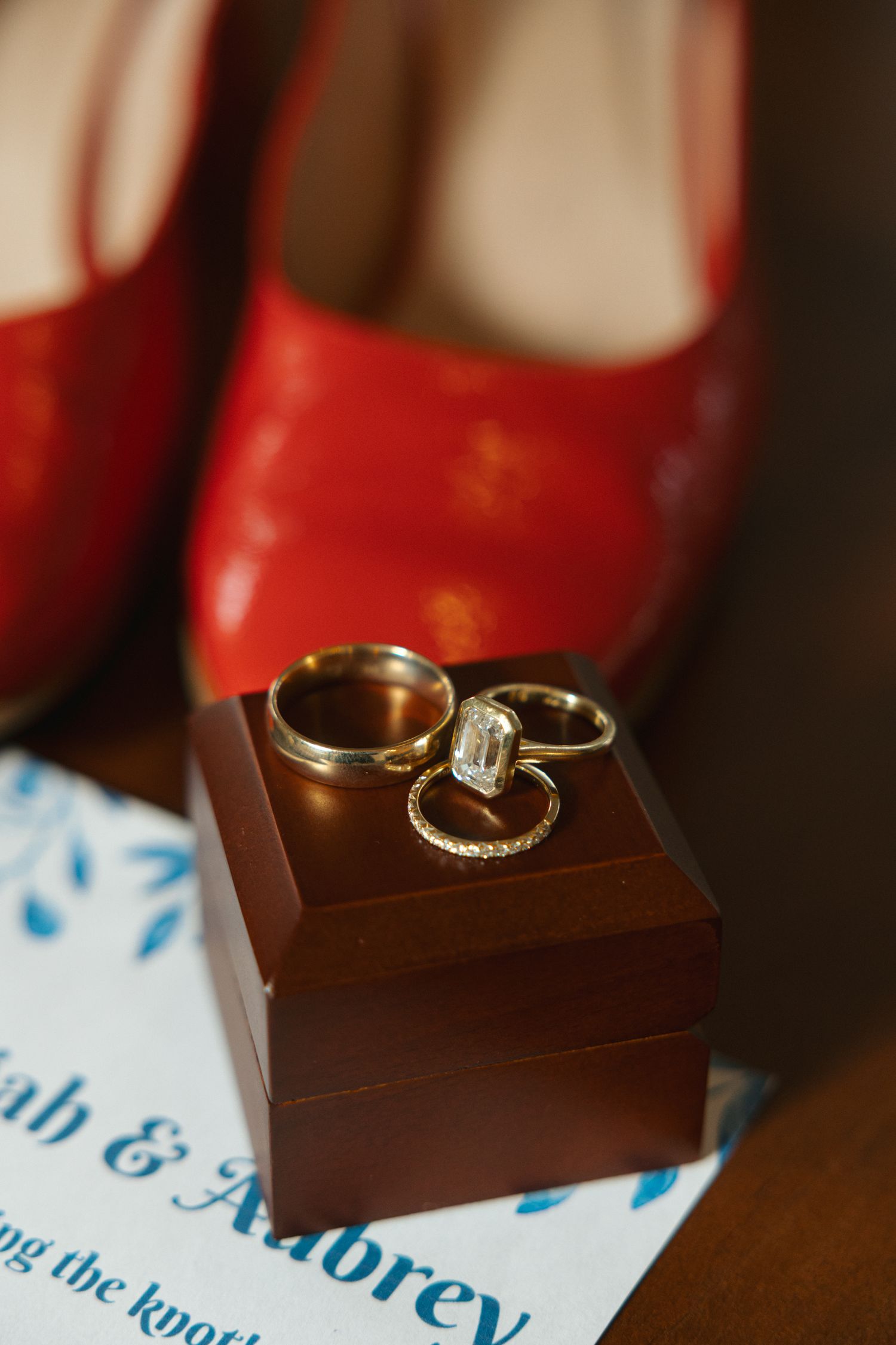 Gold wedding rings with diamond displayed on wooden box beside red heels and wedding invitation for Sarah and Aubrey.