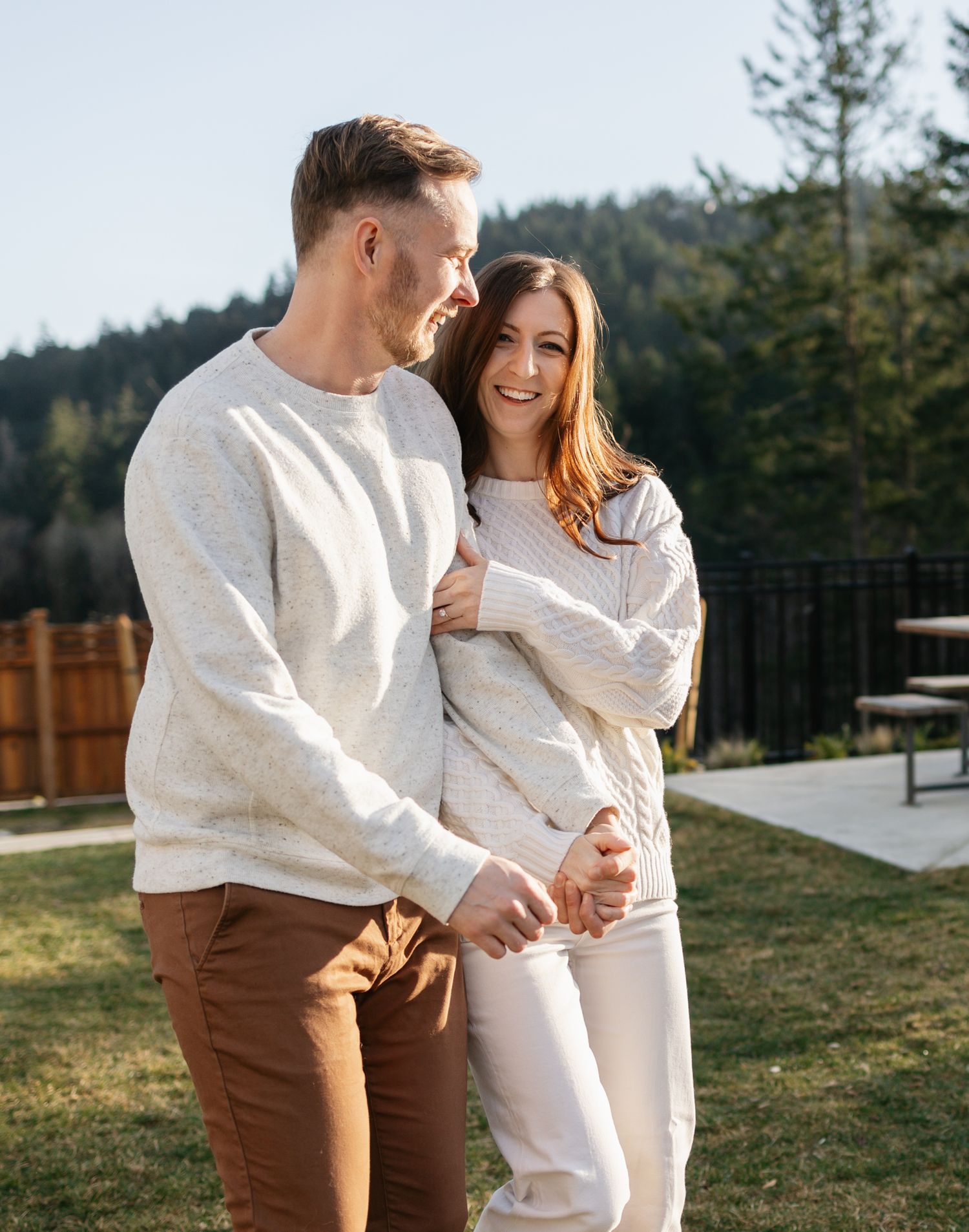 A couple in matching white sweaters and brown pants share tender moments together outdoors during a sunny day.