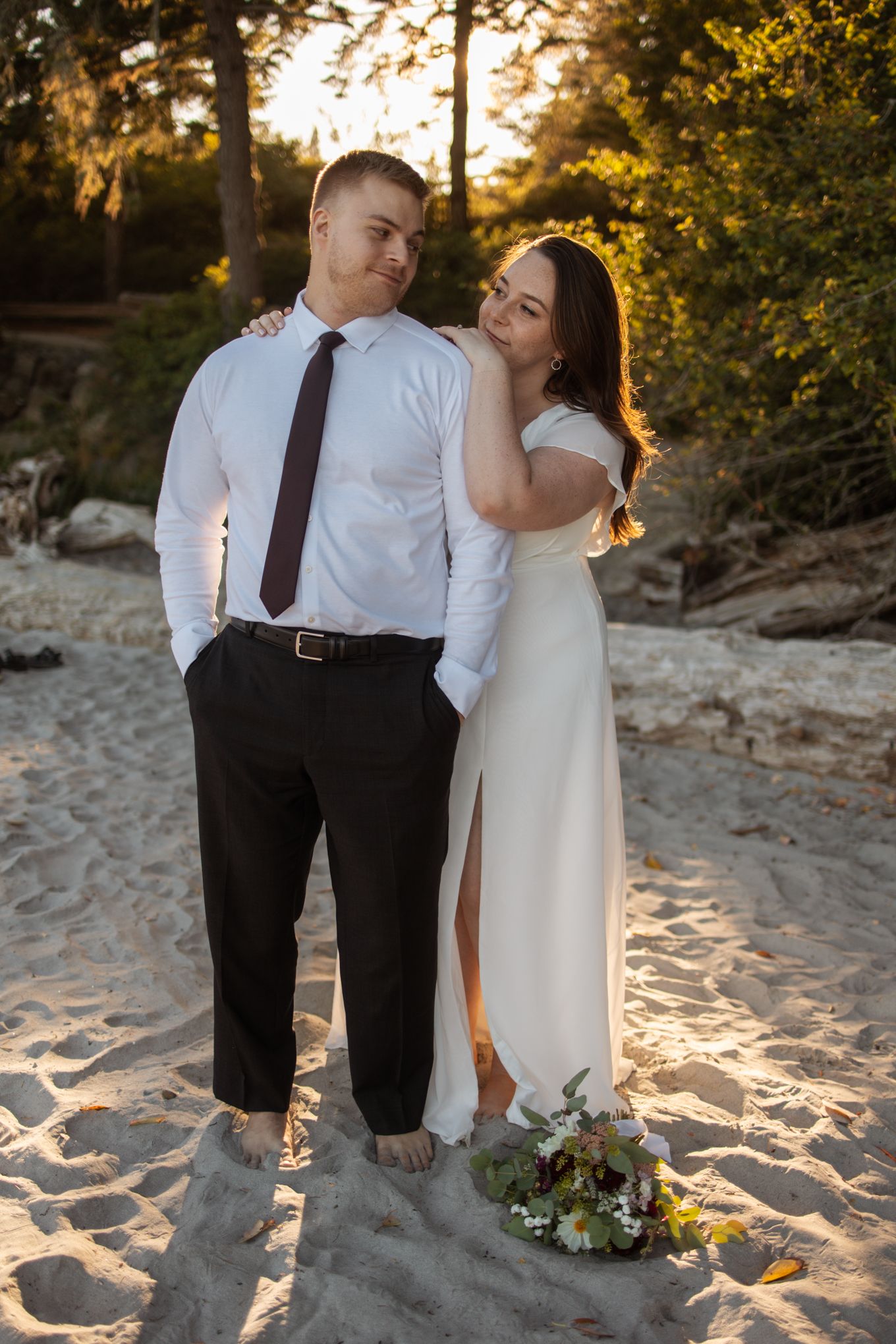 Romantic beach wedding couple embracing at sunset with sand and trees in the background.