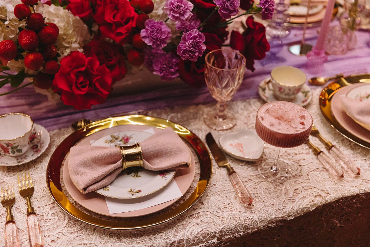 Elegant table setting with gold charger plates, pink napkins, and red and purple floral centerpiece arrangements.
