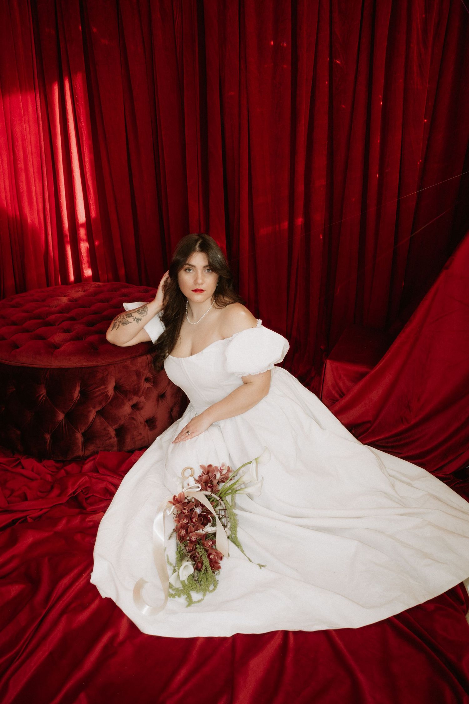 A person in a flowing white wedding dress poses dramatically against luxurious red velvet curtains with a burgundy bouquet.