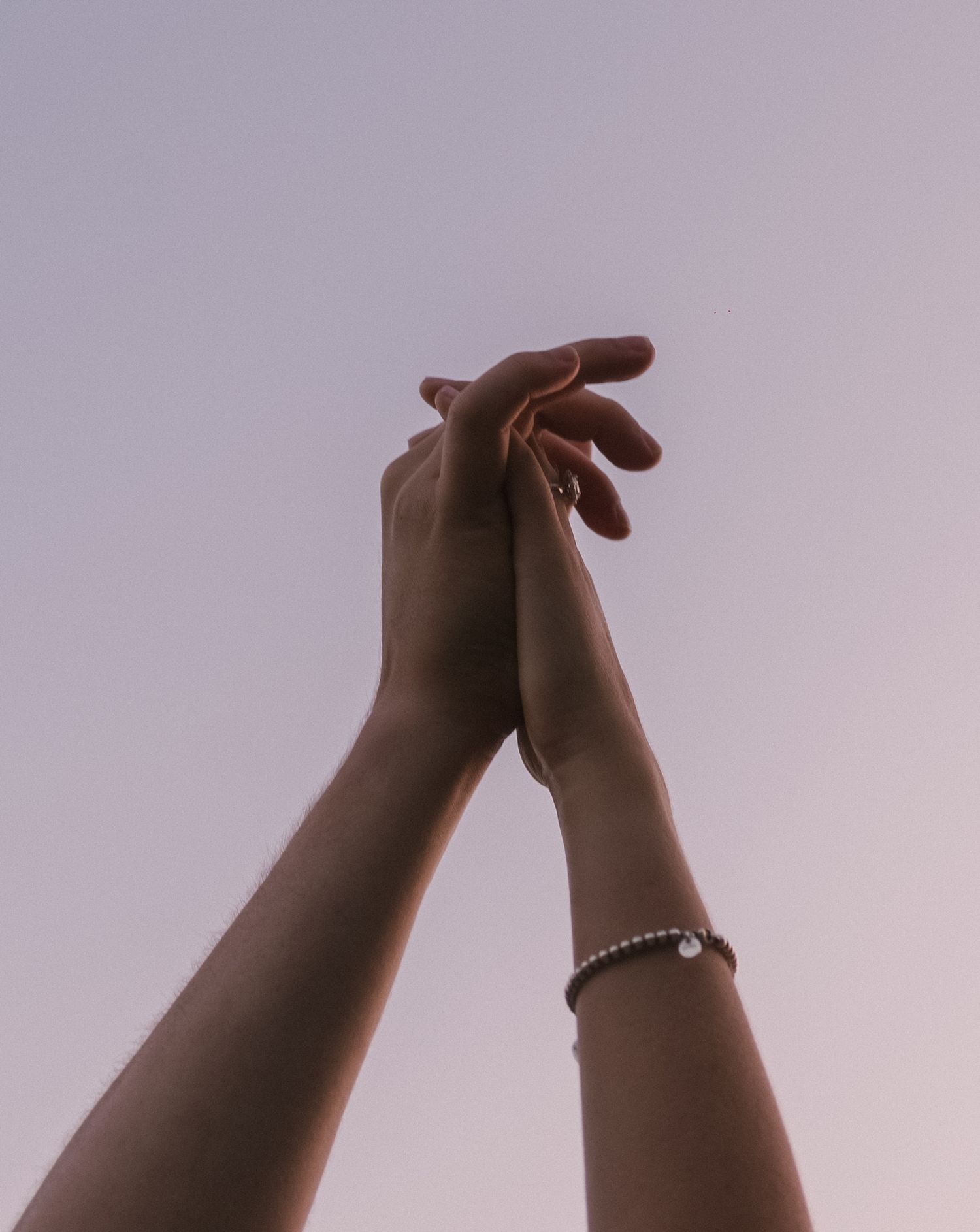 Two raised hands with a bracelet reaching up against a soft purple sky in an artistic pose.